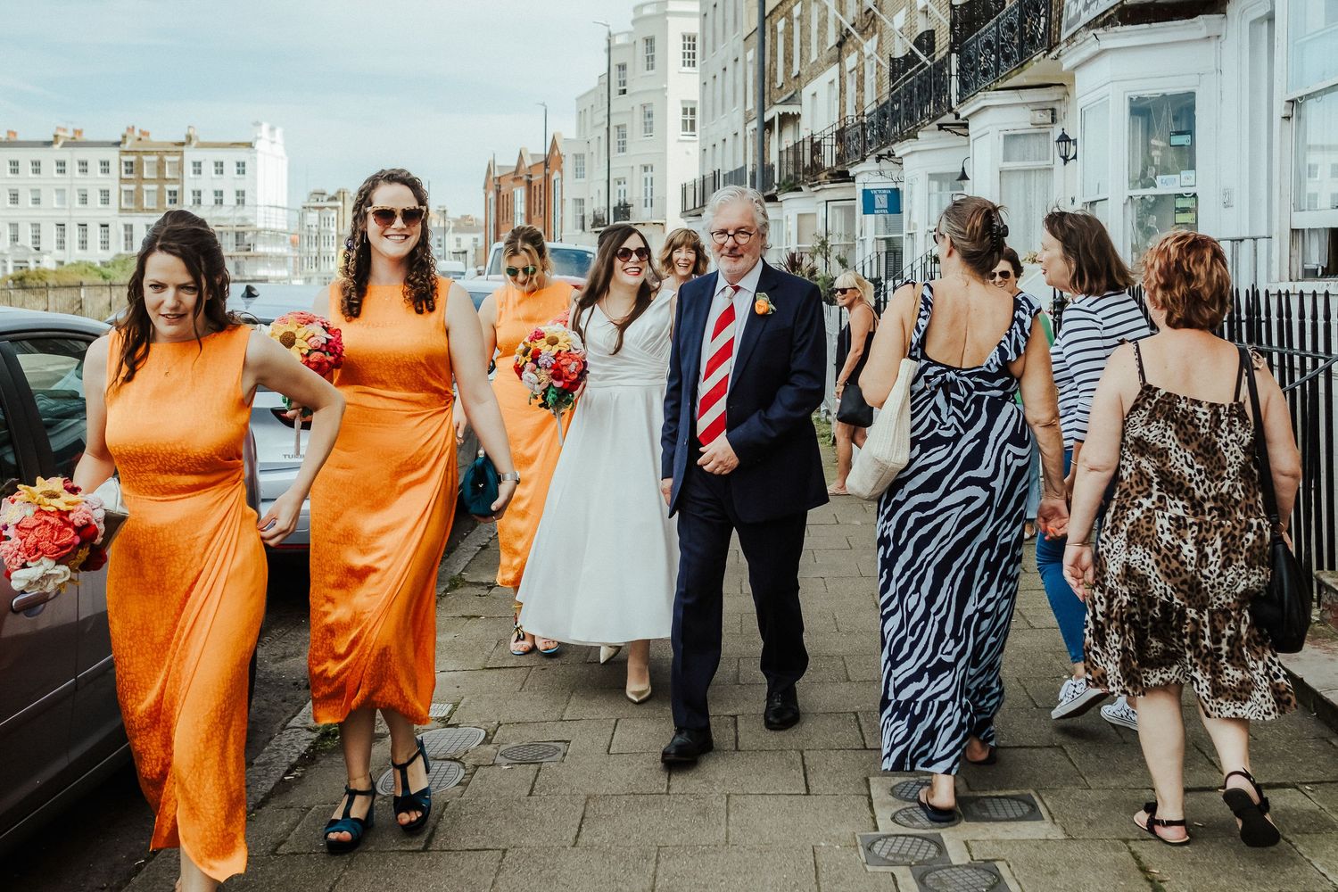 Wedding party walks along Brighton seafront with bridesmaids in vibrant orange dresses.