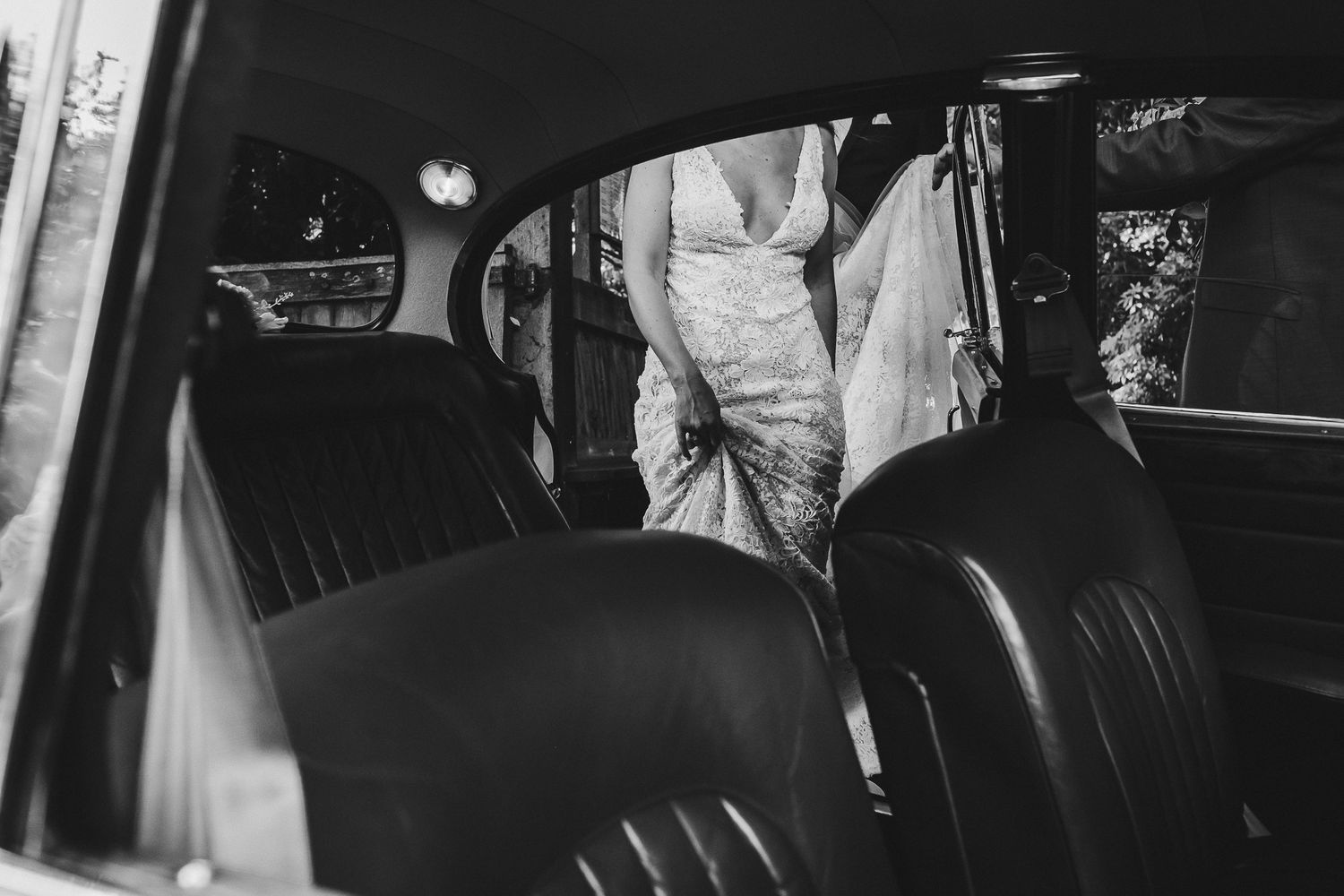 Black and white photo of a bride entering a vintage car with leather seats.