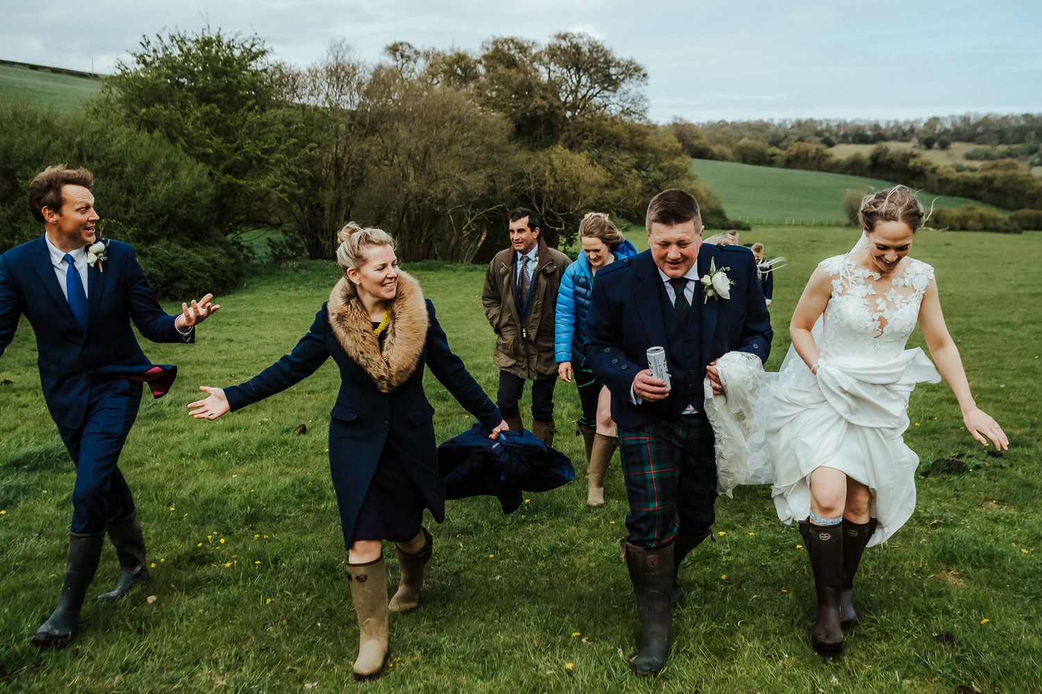 Wedding party running and laughing together across a green hillside in casual boots and formal attire.