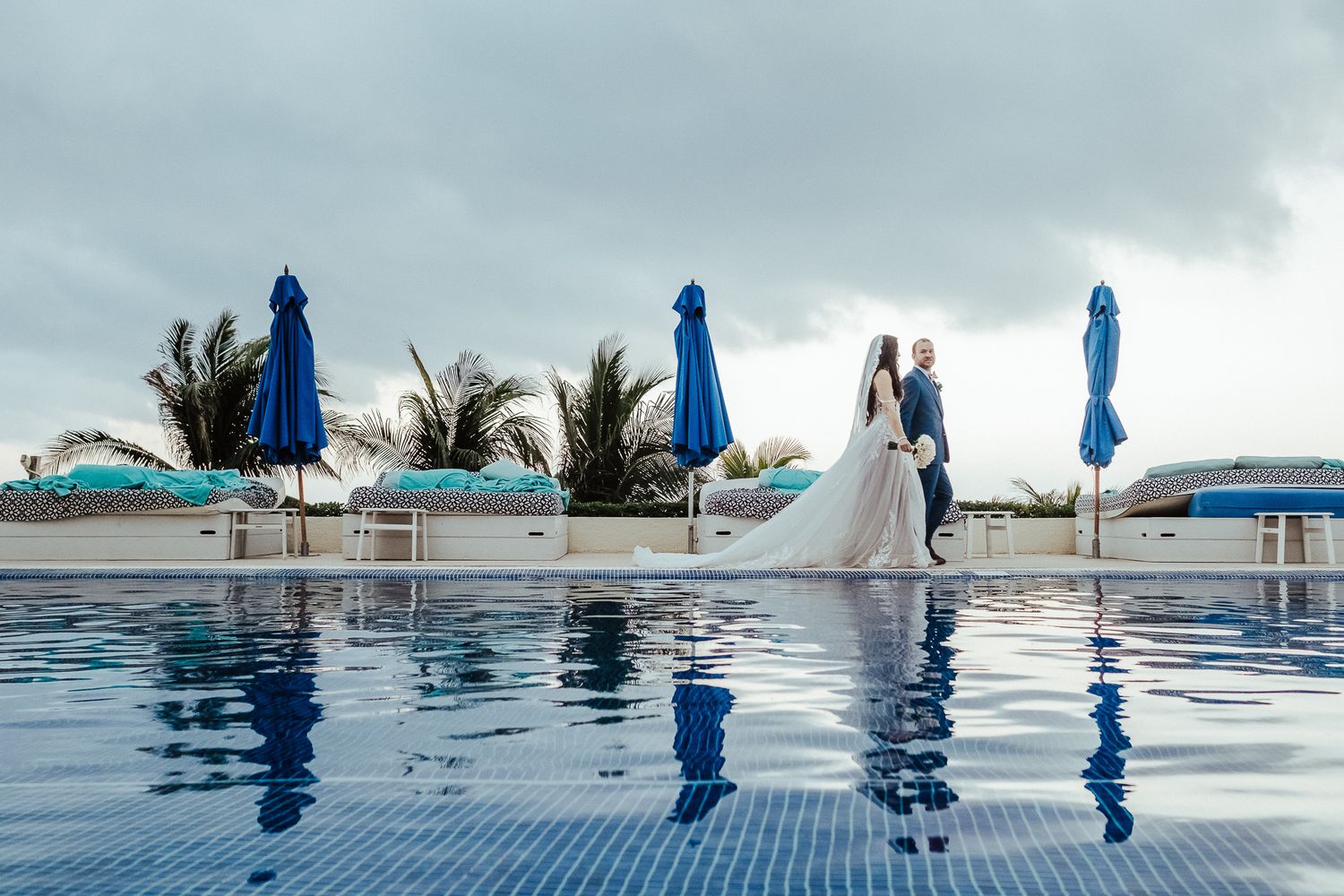 Wedding couple poses by infinity pool with blue umbrellas and ocean view.
