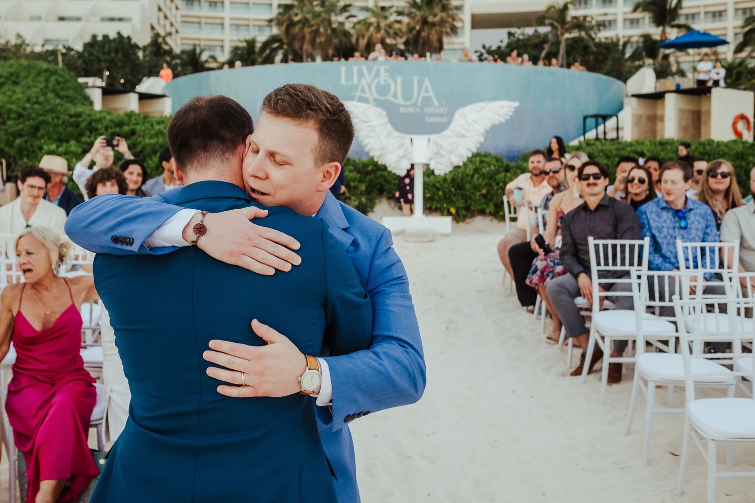 Two men in blue suits embrace during an emotional wedding ceremony moment.