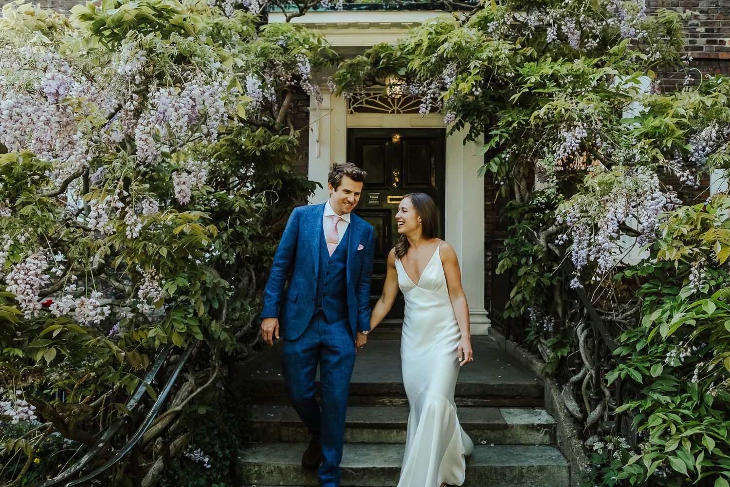 A couple descends stone steps surrounded by blooming wisteria at their garden wedding venue.