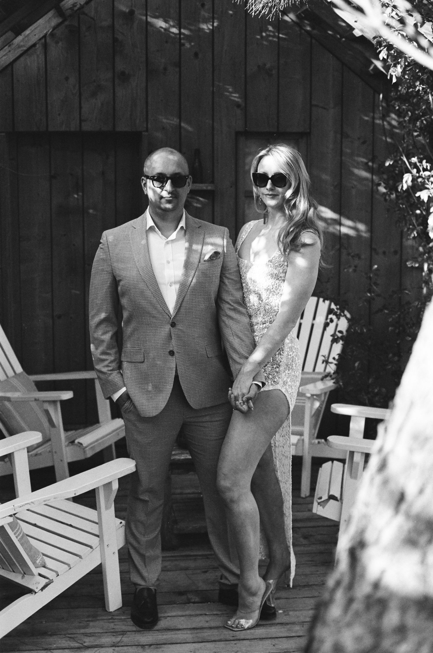 A black and white photograph of a couple posing together in formal attire on a wooden deck next to Adirondack chairs.