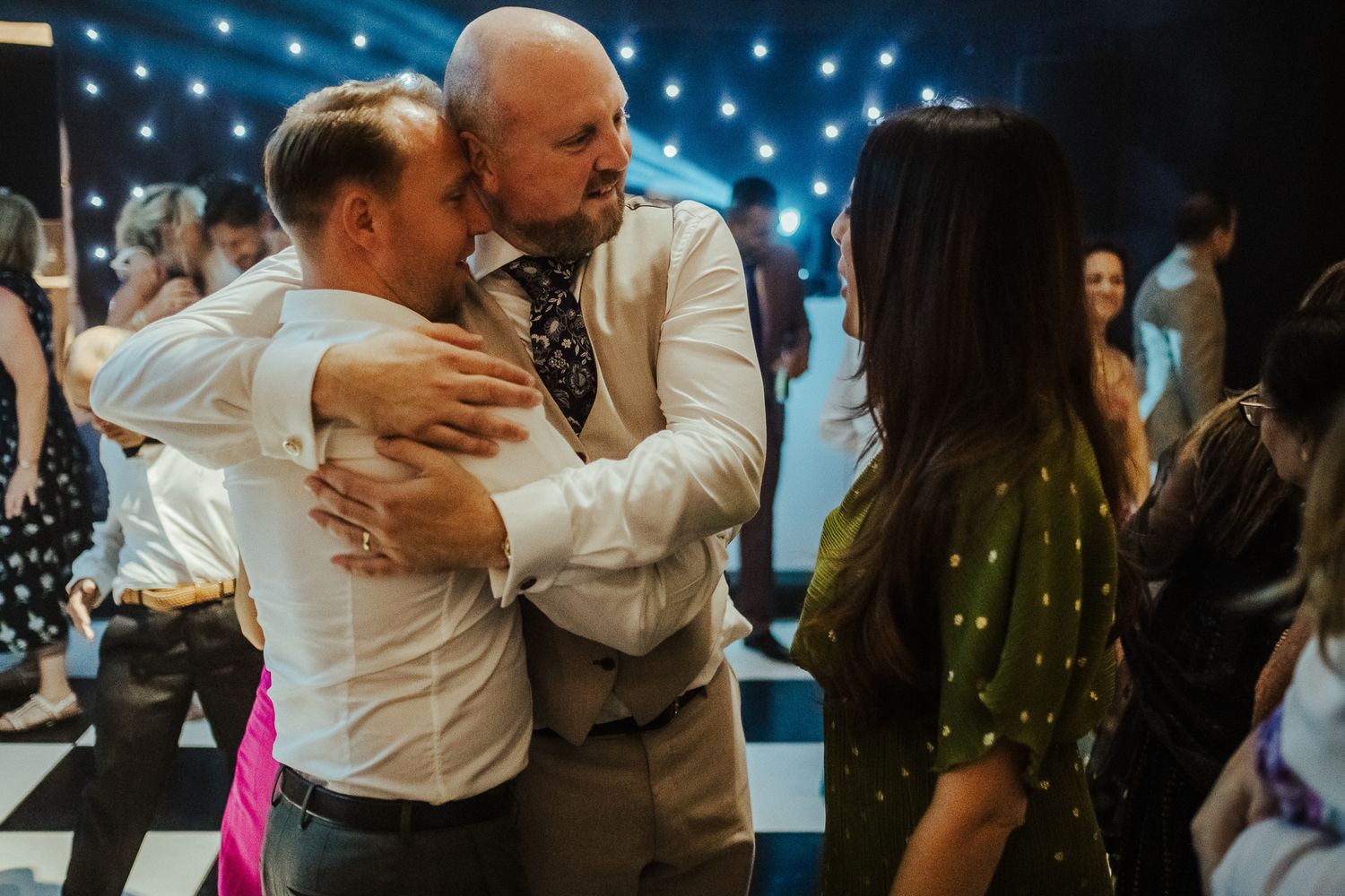 Two people share an emotional embrace while dancing on a checkered floor with string lights overhead at an indoor event.