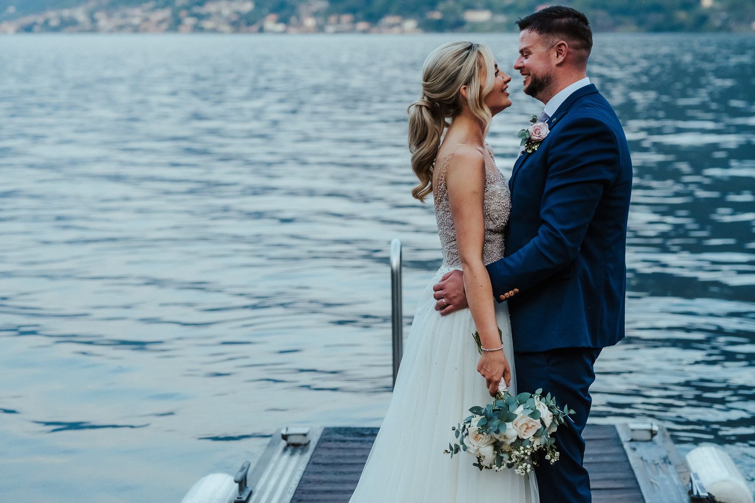 A wedding couple stands on a dock overlooking a serene lake with mountains in the background.