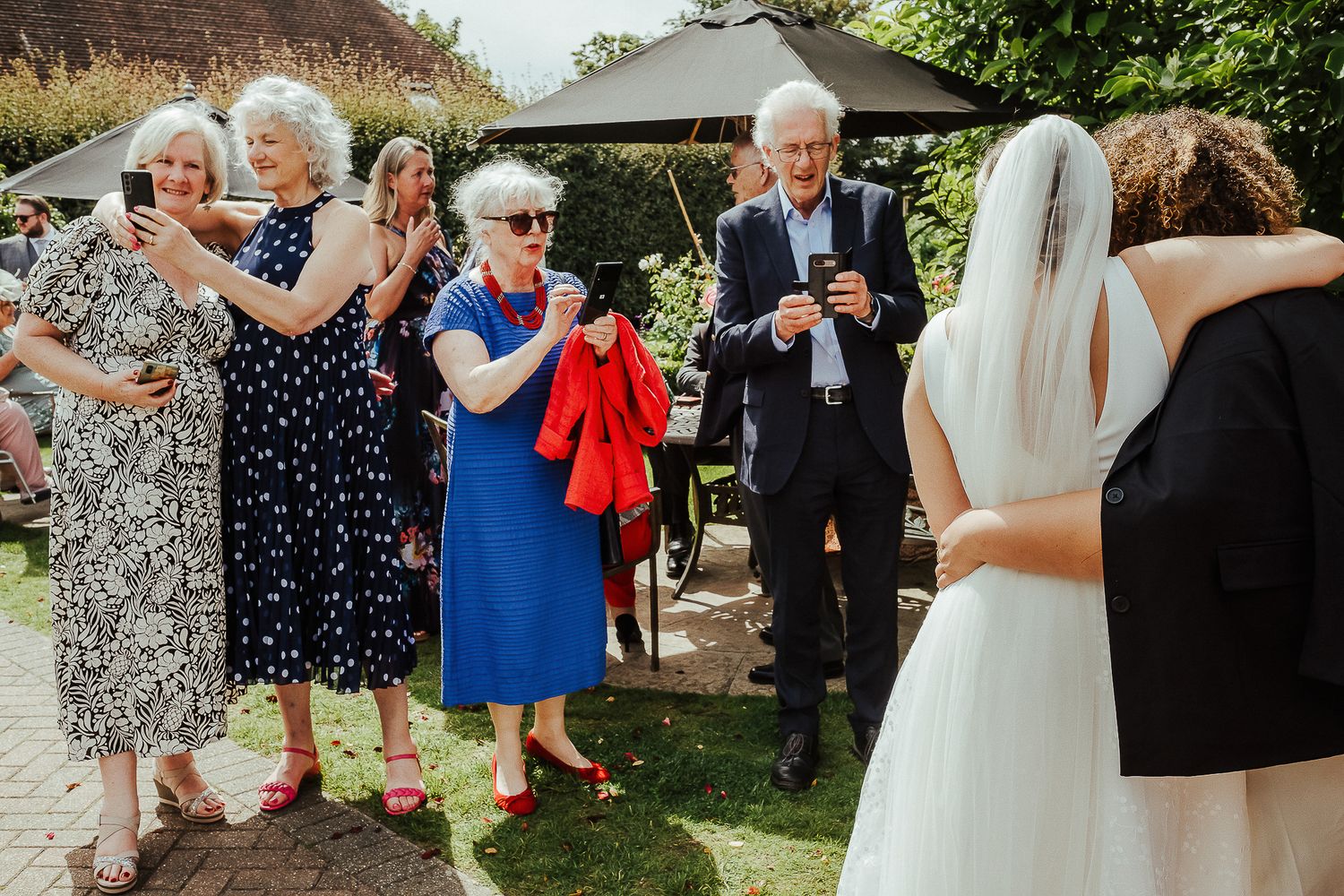 Wedding guests celebrate outdoors under a garden umbrella during a sunny afternoon reception.