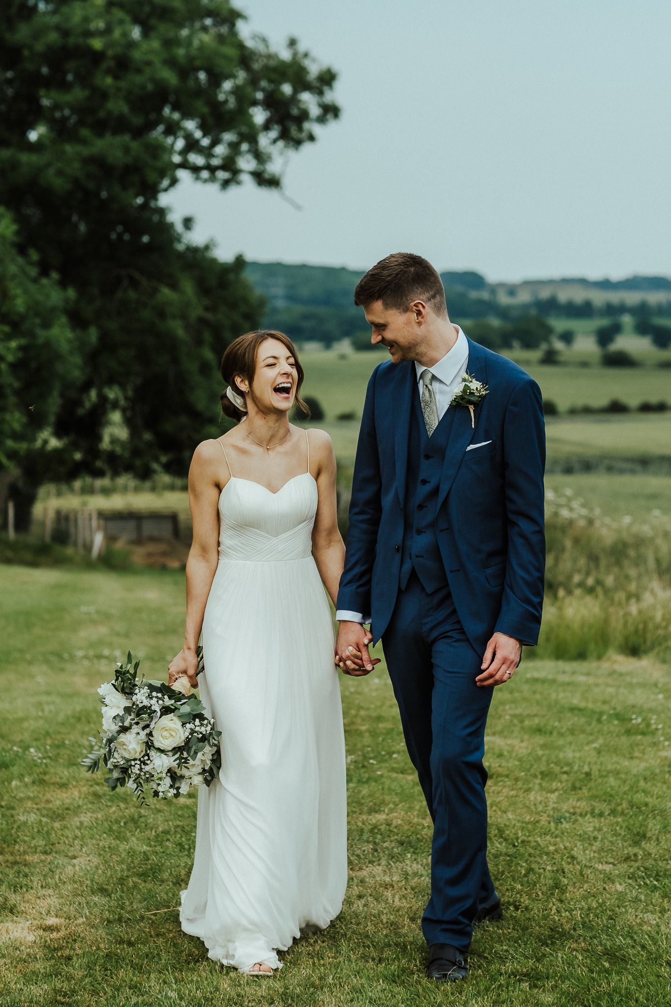 Couple shares laughs while walking on grassy field during outdoor wedding photoshoot.