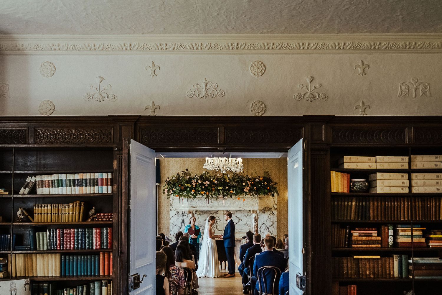Indoor wedding ceremony in a library venue with bookshelves and floral arch decorations.