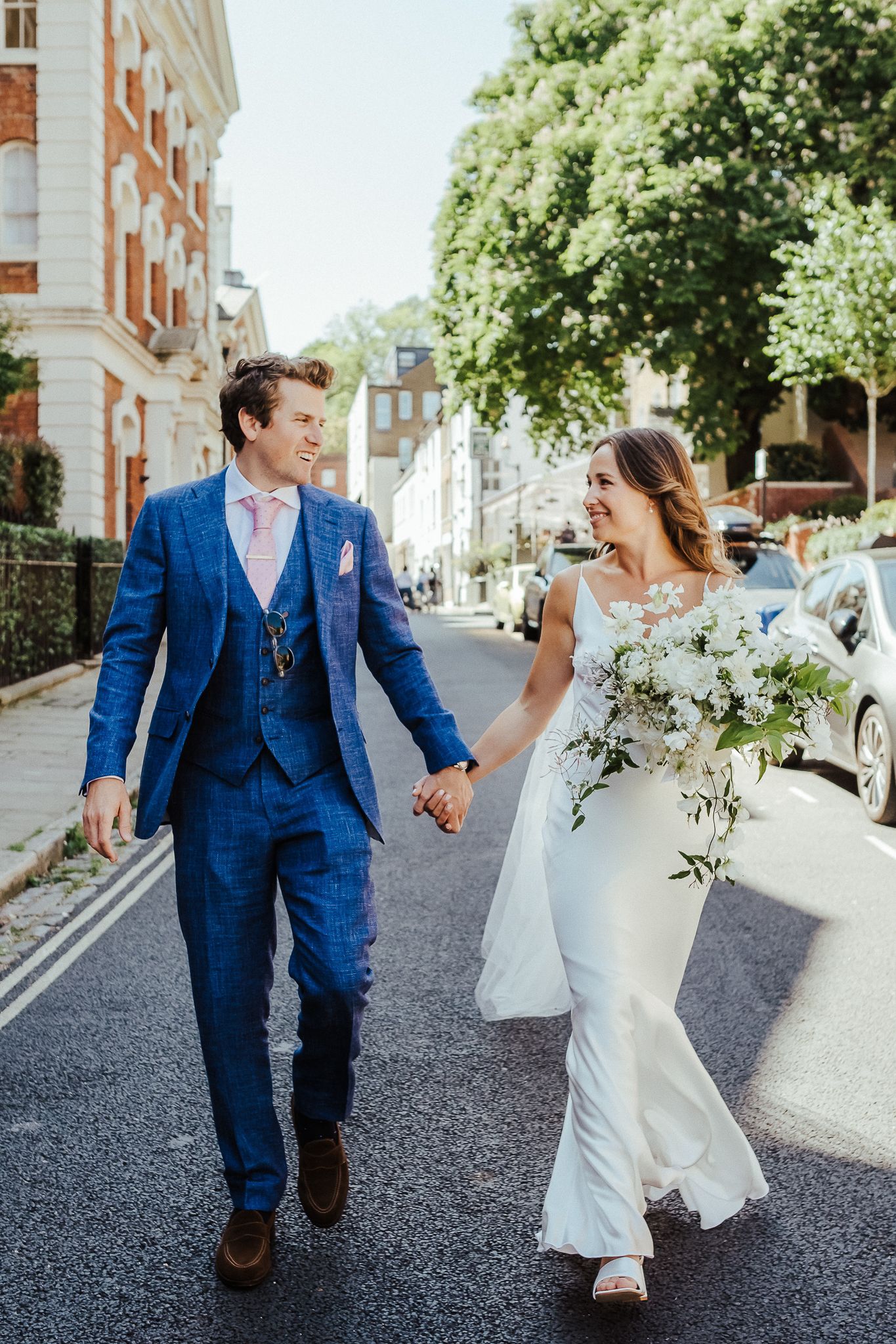 A couple walks hand in hand down a sunny London street after their wedding ceremony.