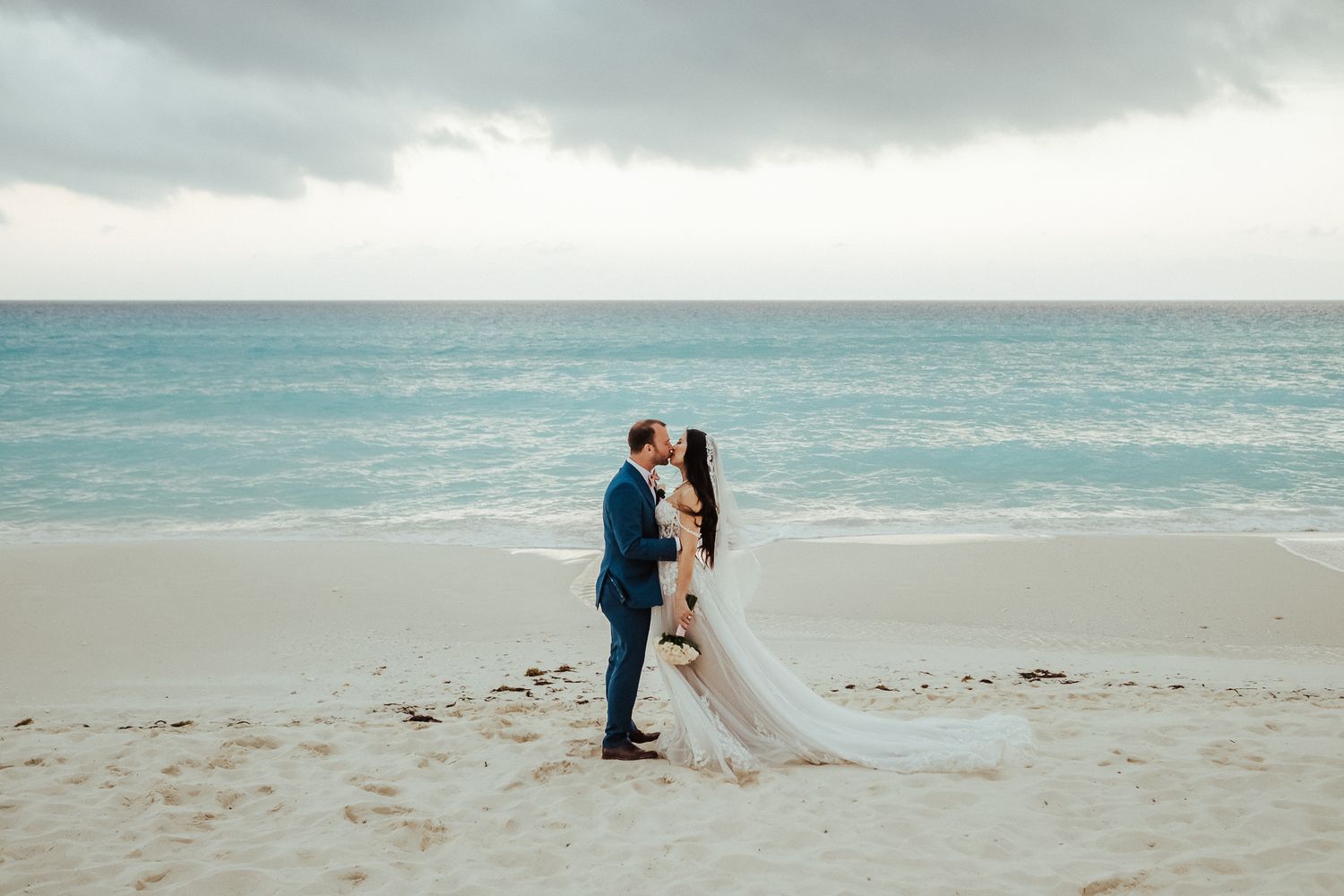 A dreamy beach wedding photo with newlyweds standing on white sand against stormy turquoise waters.