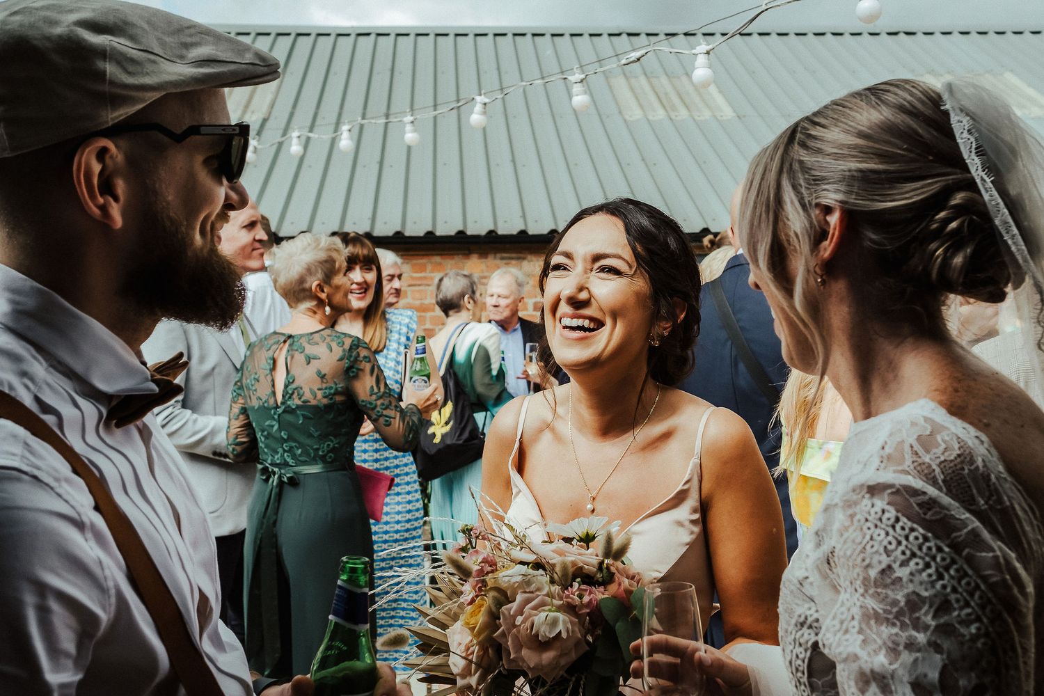 Wedding guests laugh and celebrate with drinks at an outdoor reception under a metal roof.