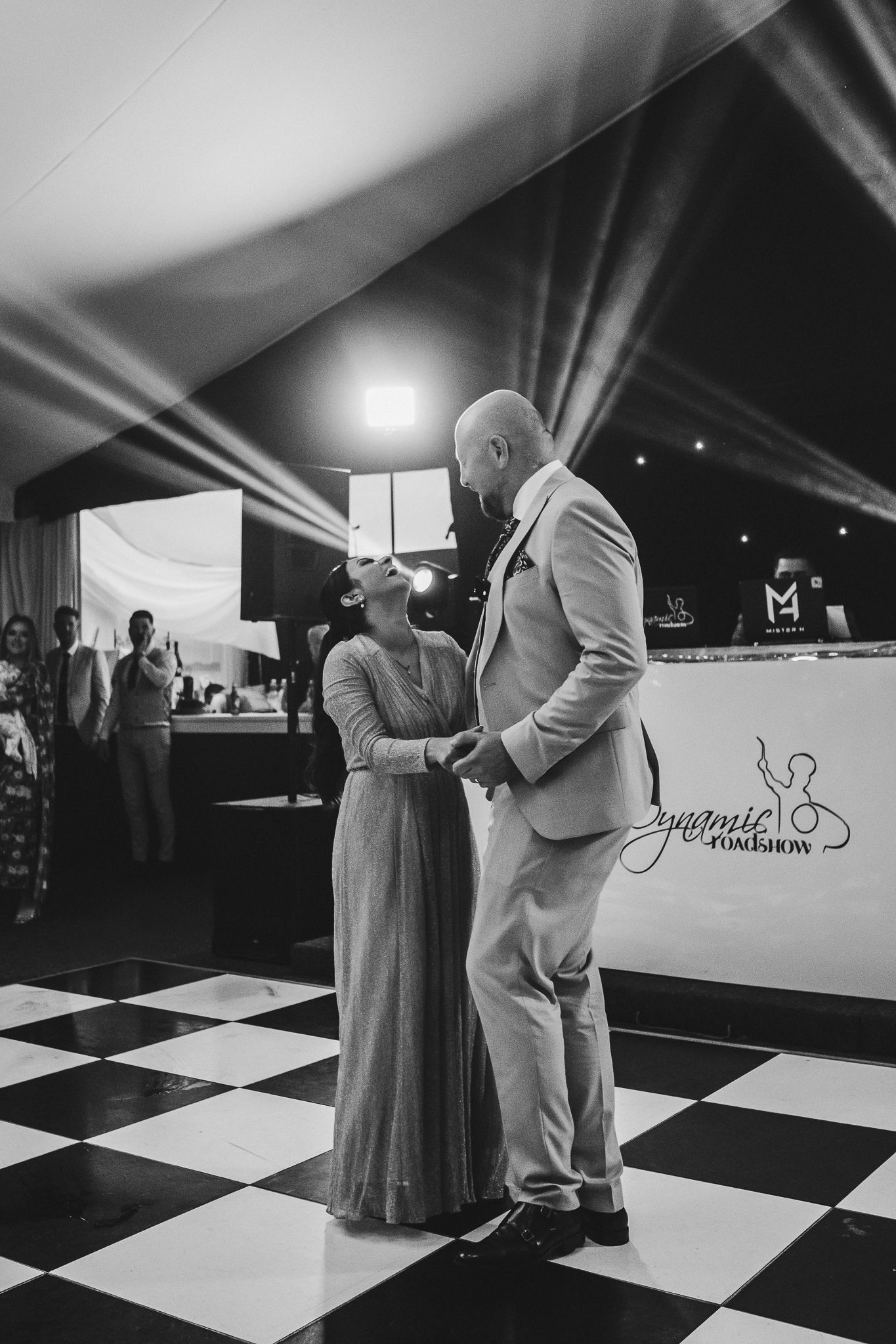Dance scene captured in black and white on a checkerboard floor during a wedding reception celebration.