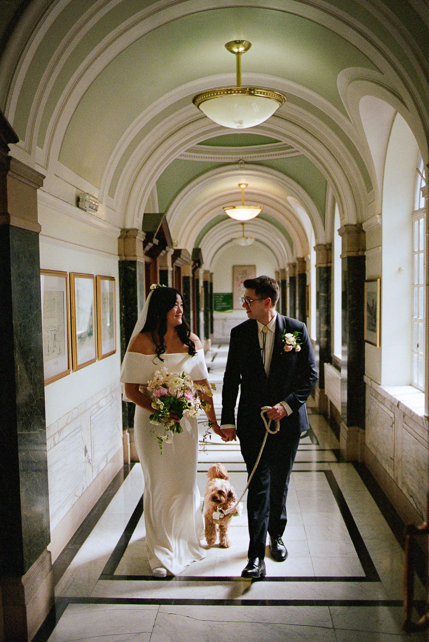 Wedding couple walks through elegant arched hallway with marble columns and architectural details in romantic pose.