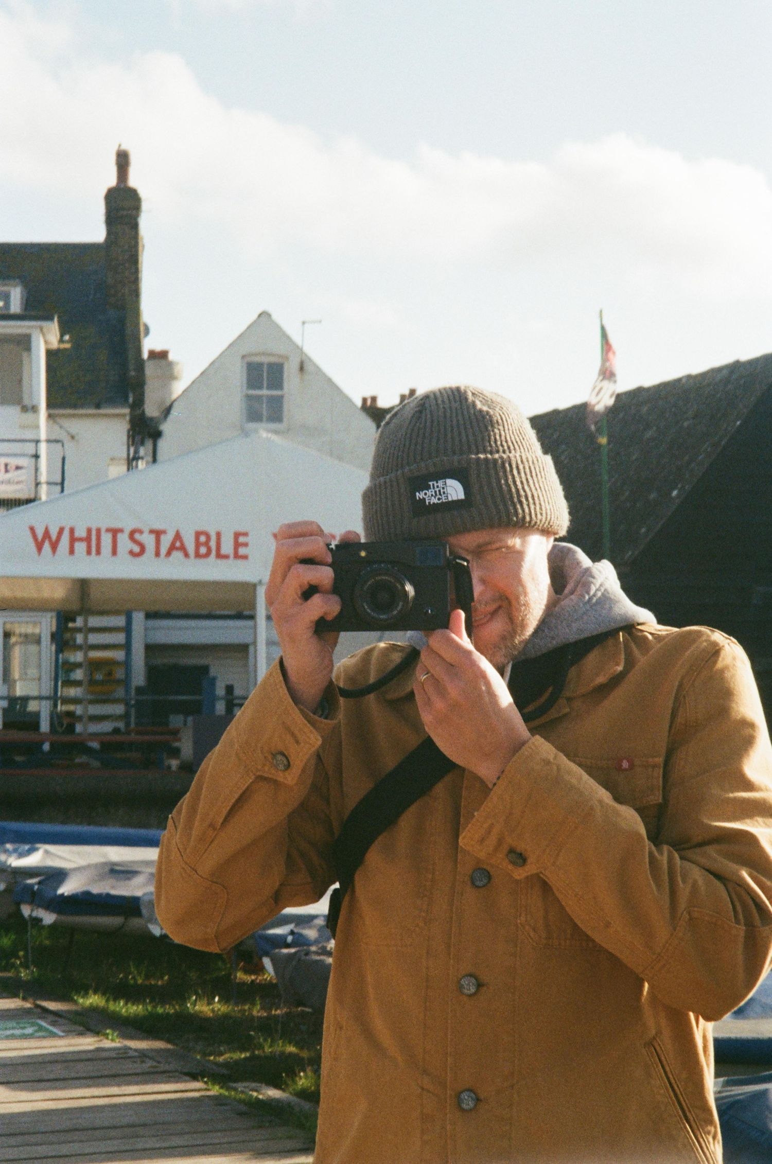 Person wearing beanie and brown jacket takes photos in front of Whitstable storefront.