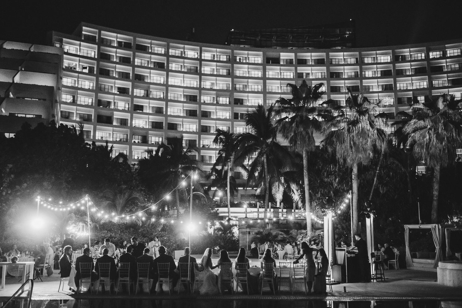 Illuminated resort hotel at night with palm trees and crowd gathered for outdoor celebration.
