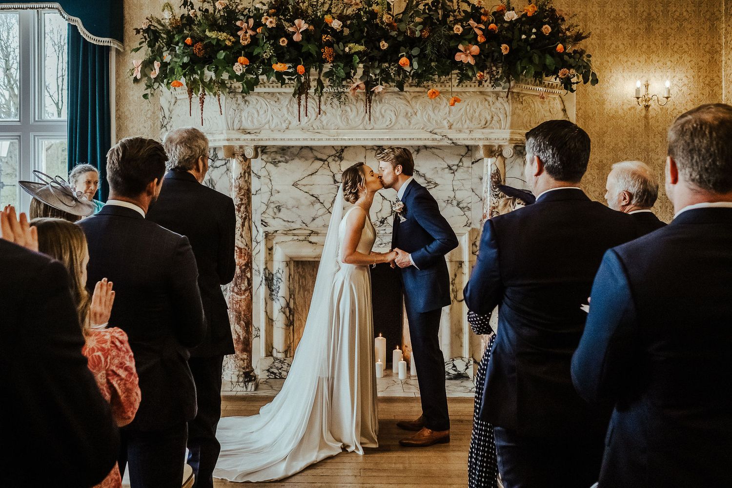 Couple sharing first kiss during wedding ceremony as guests look on in elegant room with floral decor.