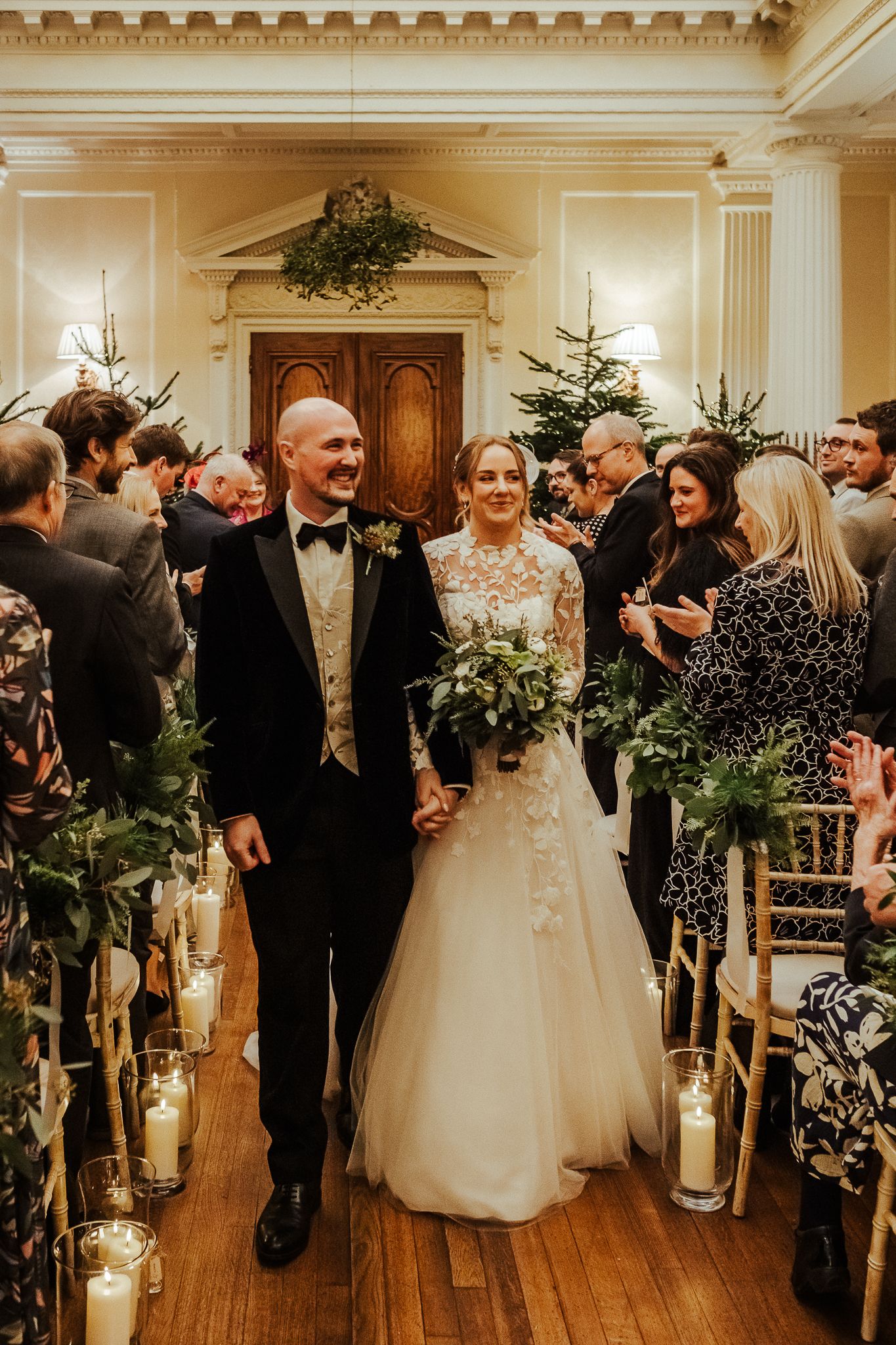 Wedding recessional in grand hall with Christmas trees and candlelit aisle.