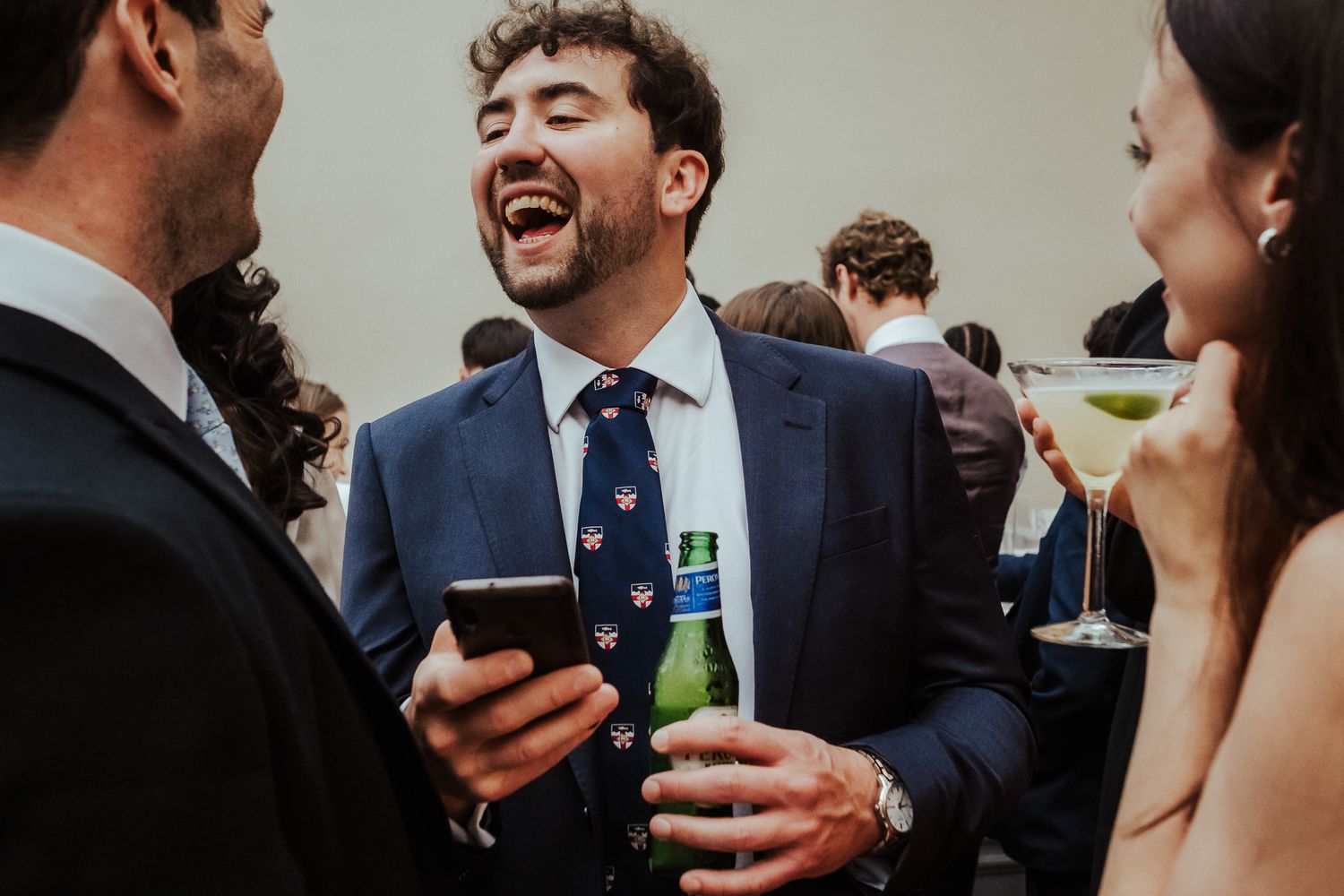 Candid shot of wedding guests laughing while sharing drinks and looking at phone.
