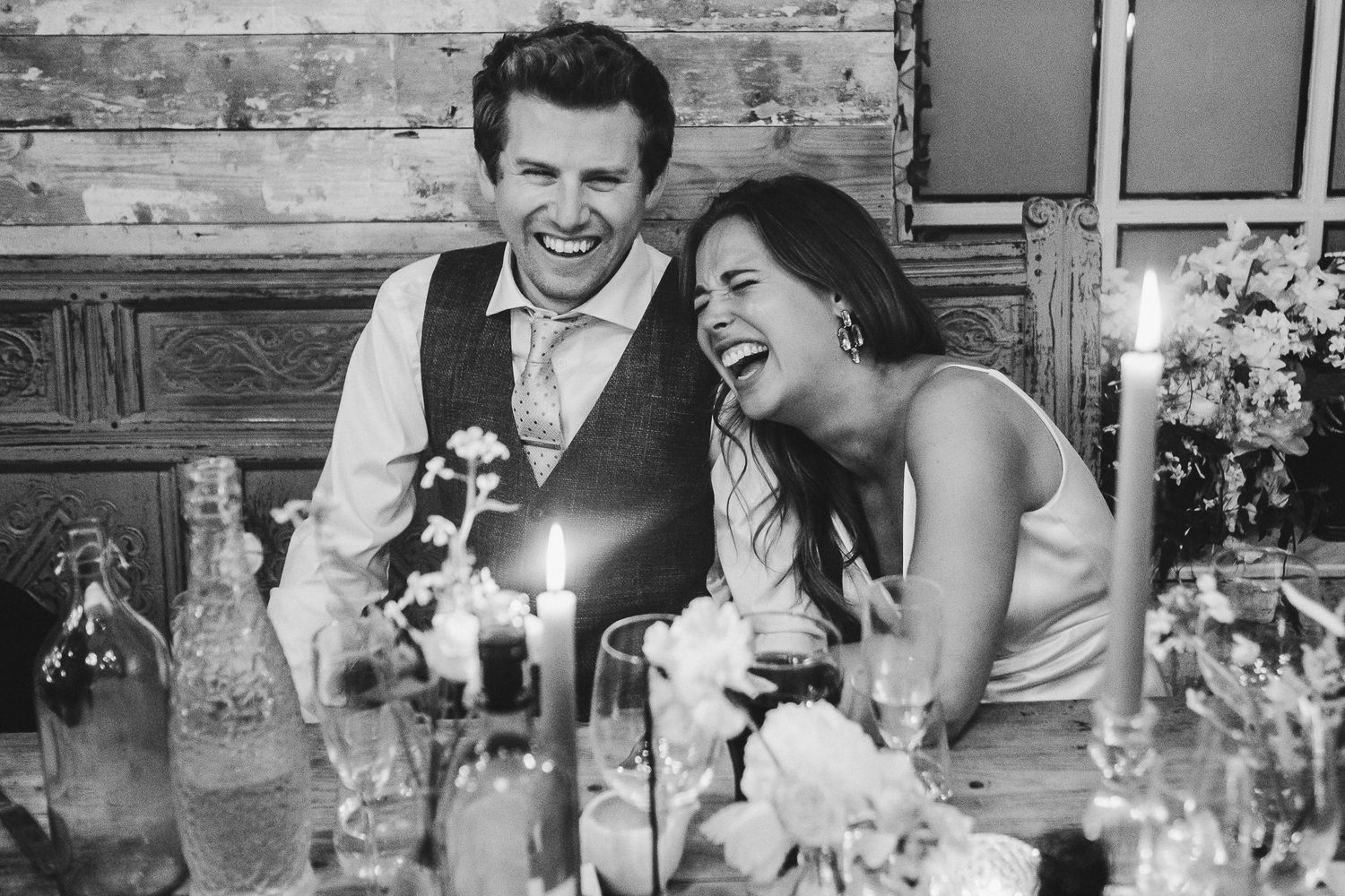 Couple shares genuine laughter at candlelit dinner table surrounded by glassware.