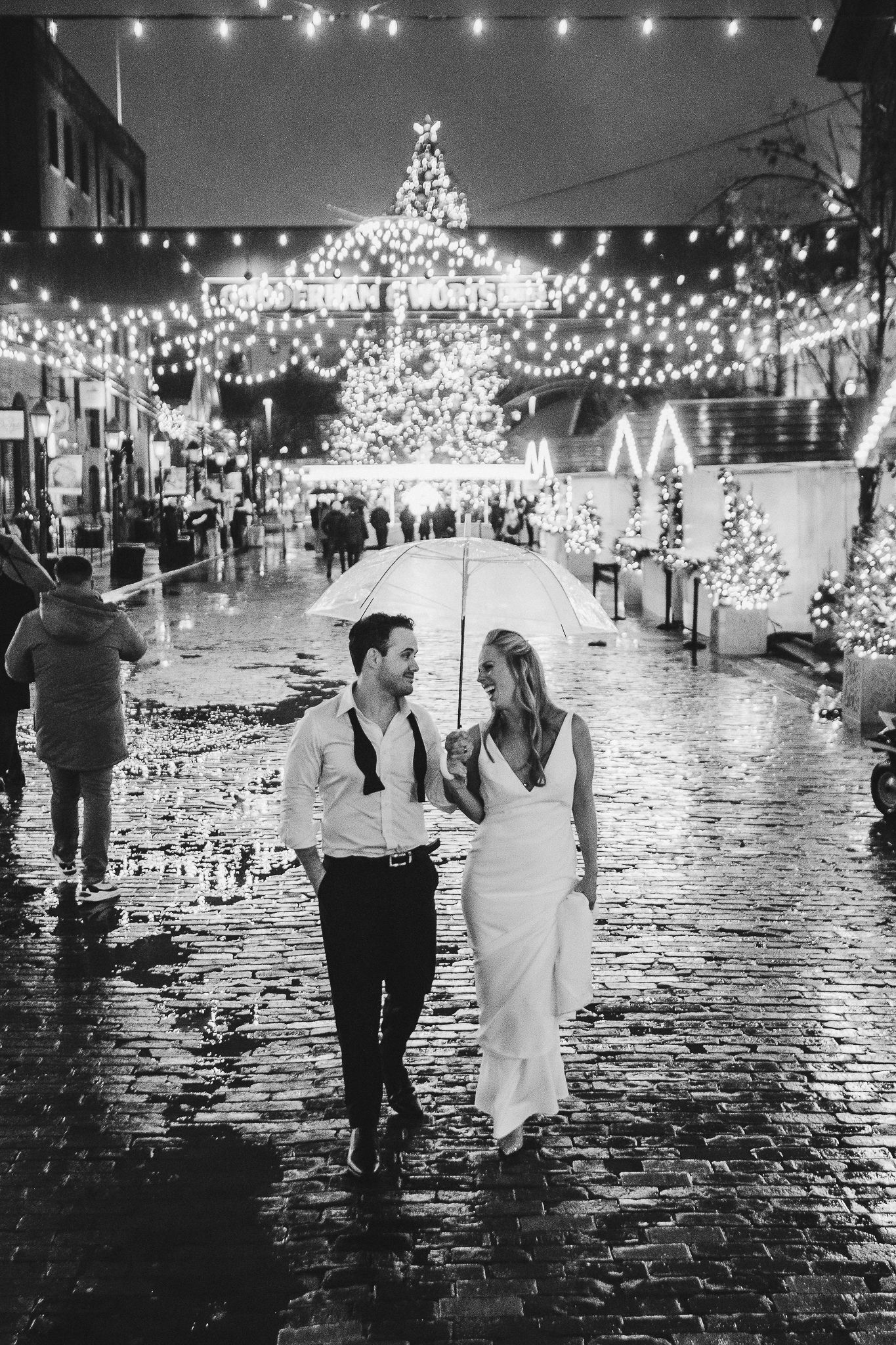 Wedding couple walks down rainy cobblestone street illuminated by string lights and glowing Christmas trees.