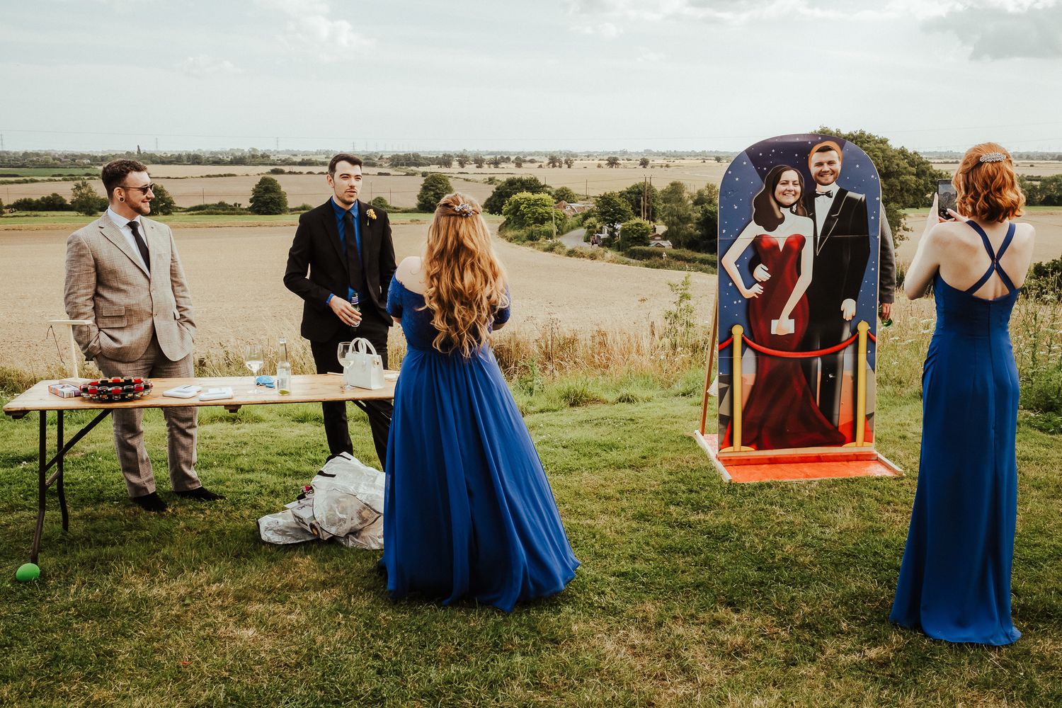 Wedding party in formal attire poses on grassy hilltop with swing set backdrop.