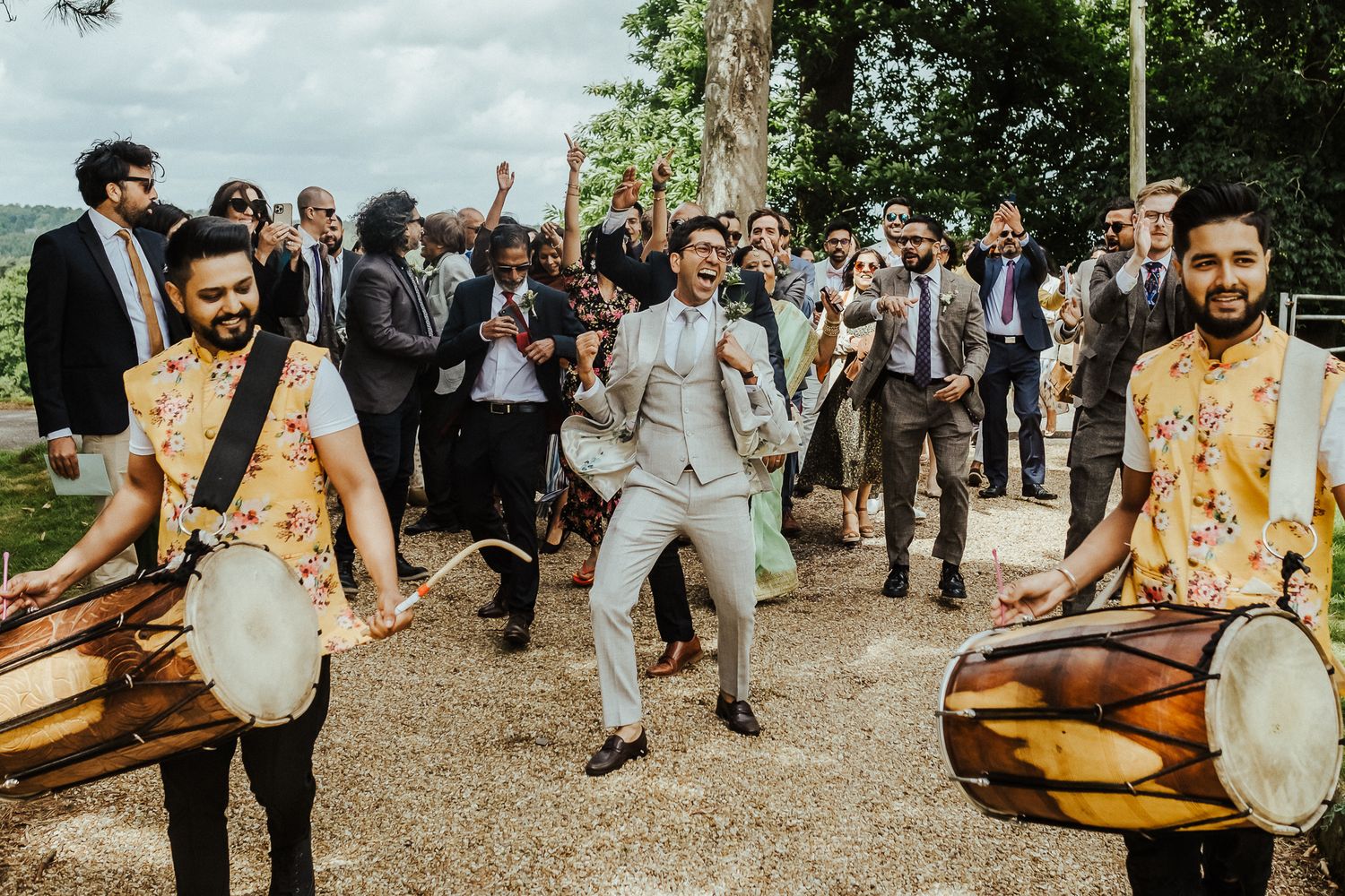 Drummers in floral shirts lead a joyful wedding procession with dancing guests following behind.