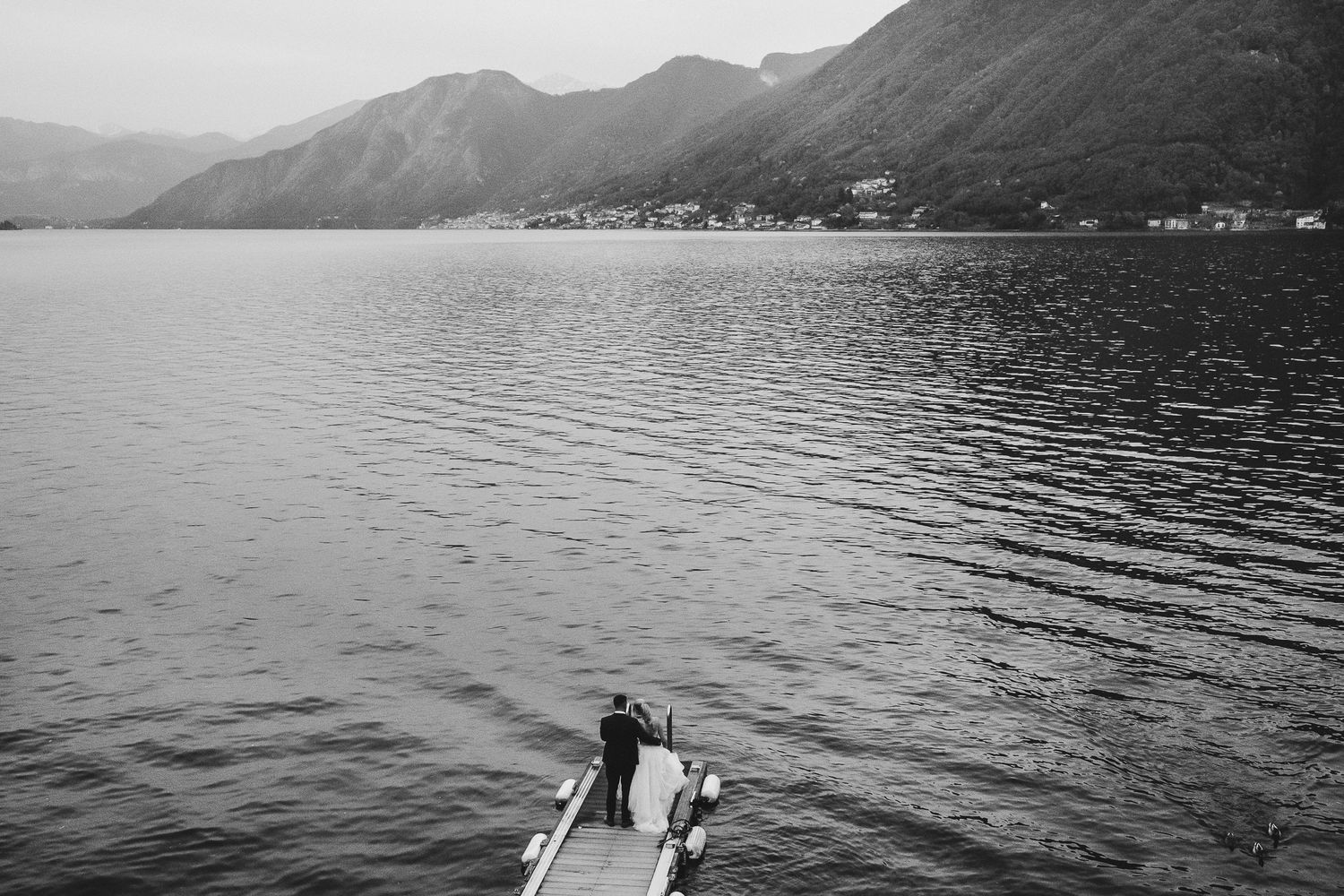 Aerial black and white view of a couple standing on a dock over a mountain lake.