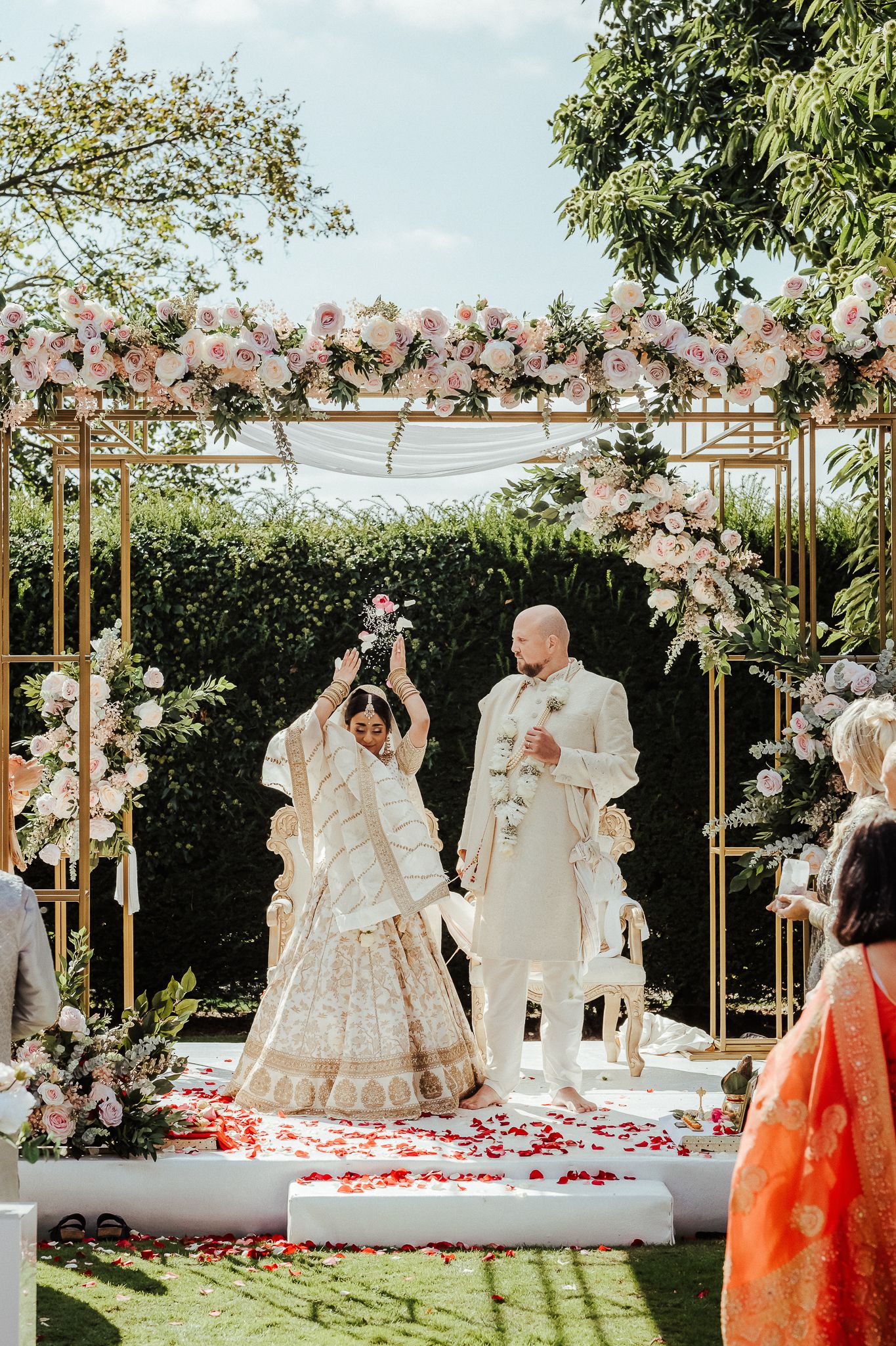 A romantic outdoor wedding ceremony setup with pink floral arrangements and wooden pergolas along a path adorned with rose petals.