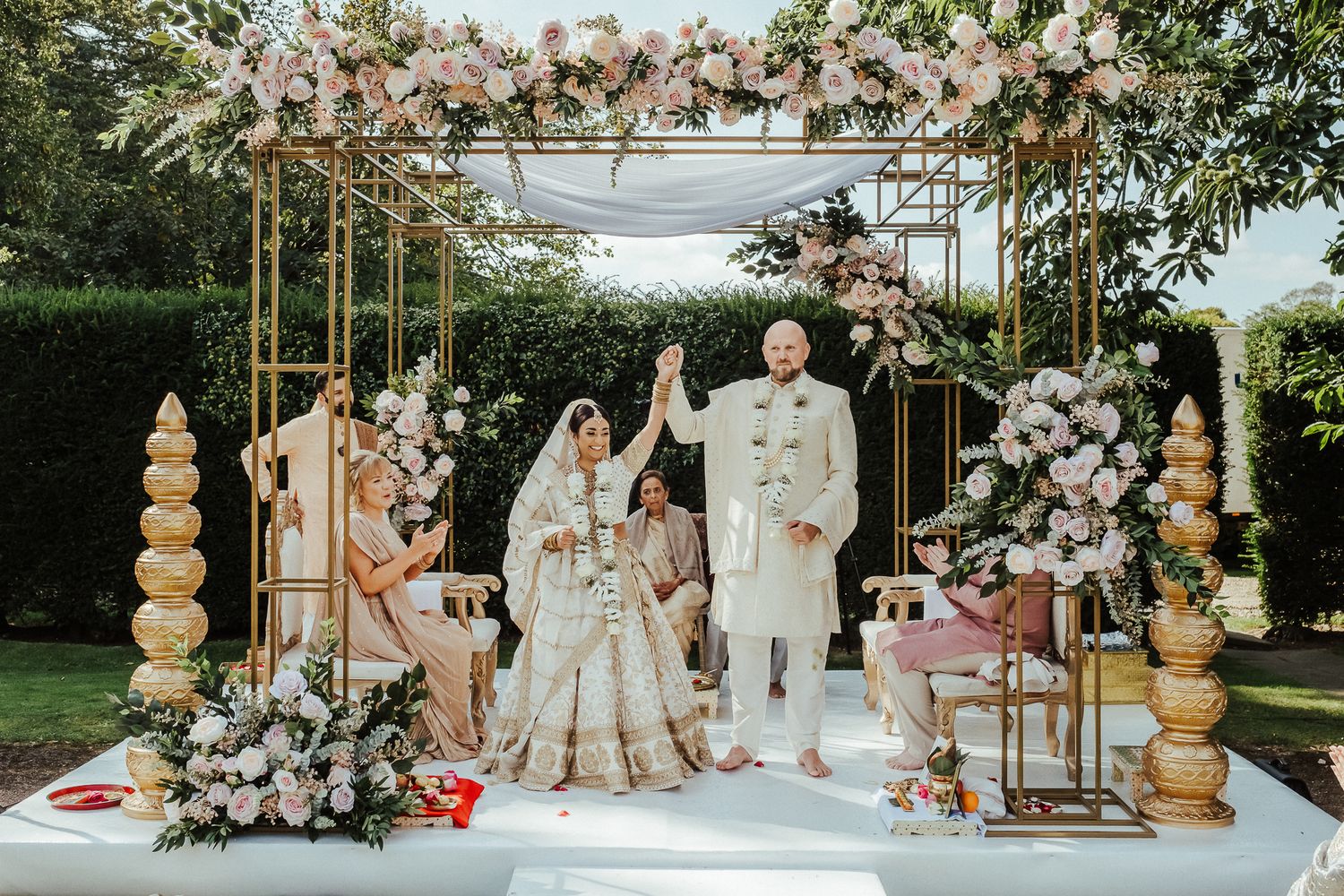 Traditional Indian wedding ceremony setup with floral arch, gold decor elements and elegant white platform in outdoor garden setting.