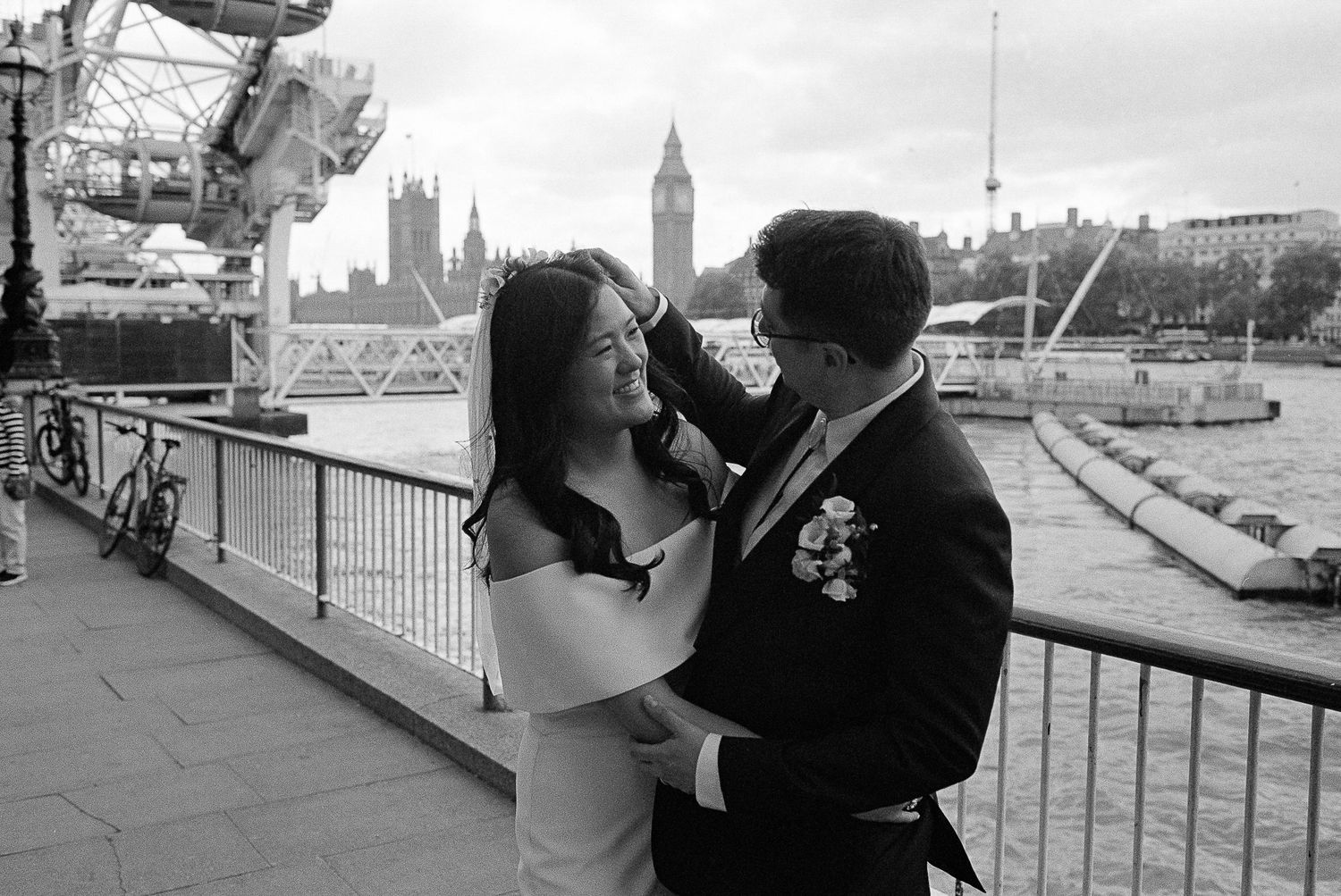 A romantic couple embrace on the South Bank of London's River Thames with Big Ben and Parliament visible in the background.