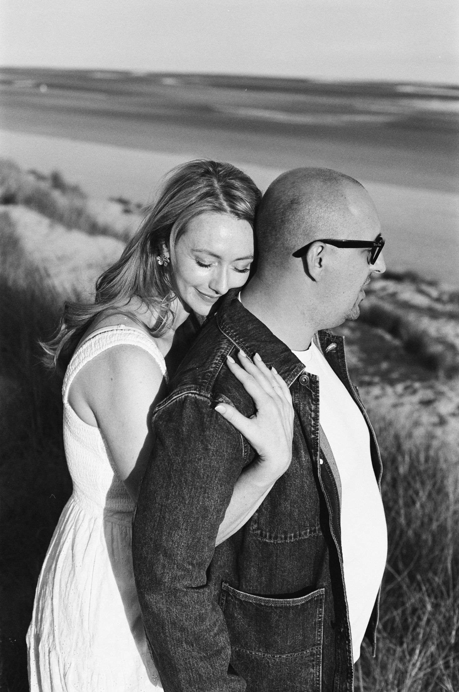 A romantic couple in a field shows tender affection as a woman in white dress leans on person wearing denim vest.