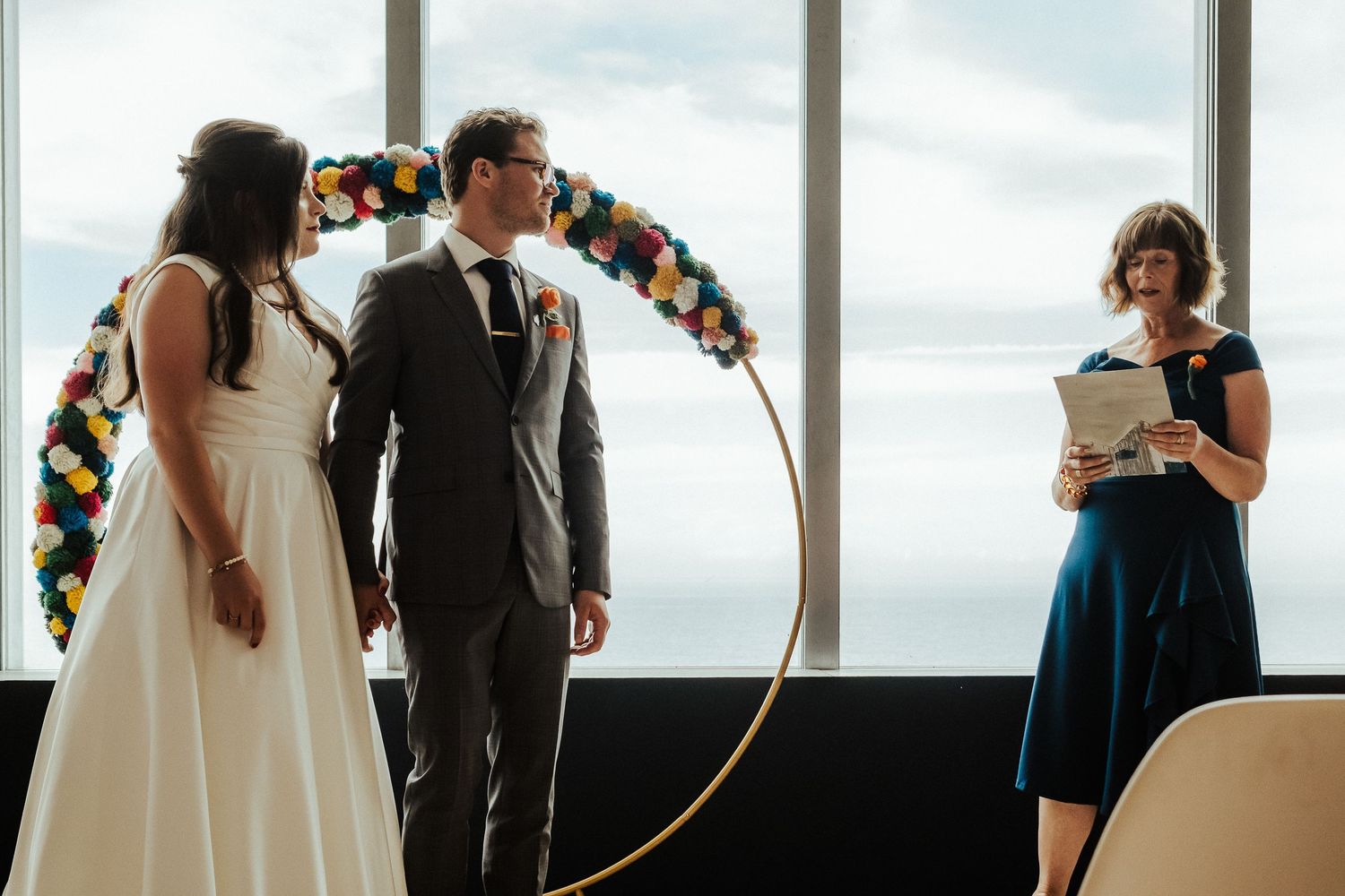 Wedding ceremony under circular balloon arch with ocean backdrop and officiant reading vows.