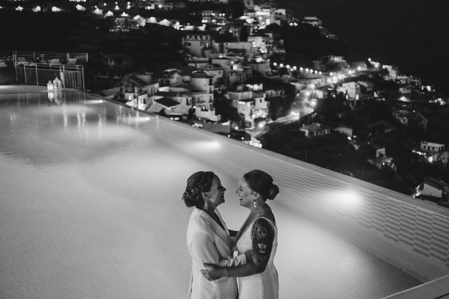 Nighttime cityscape view of Amalfi Coast with twinkling lights reflecting on water.