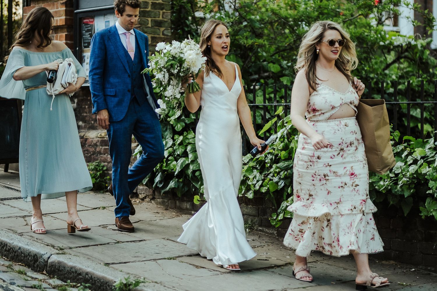 A wedding party walks down a garden path with bridesmaids in flowing dresses.