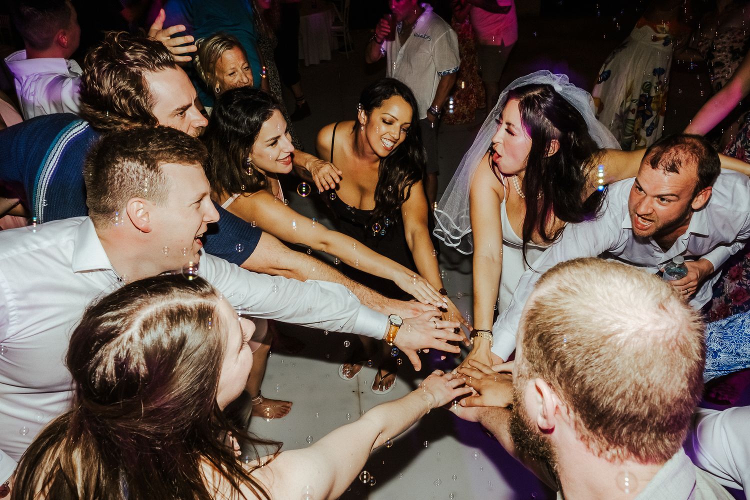 A joyous crowd reaches their hands inward in a circle during a wedding reception dance party celebration.