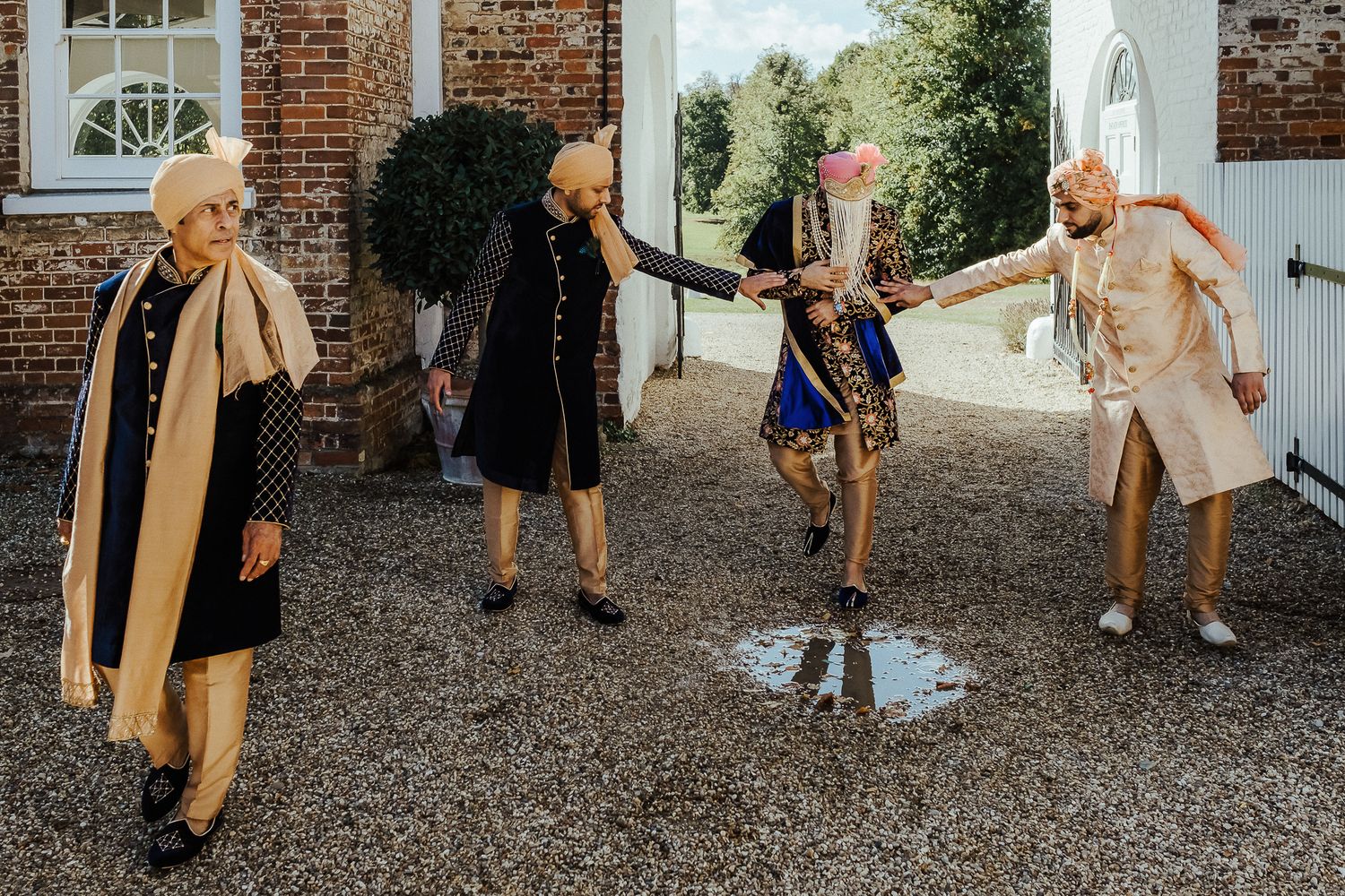 Four people in long coats dance on a gravel path in front of a brick building.