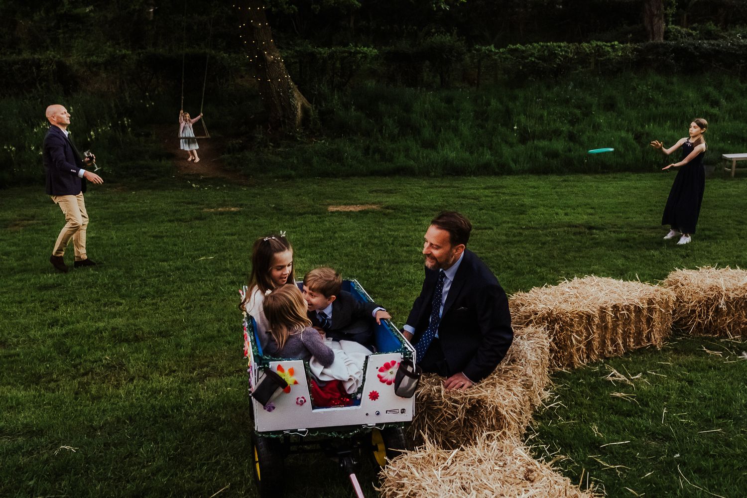 Wedding guests relax on hay bales at an outdoor rustic countryside reception.