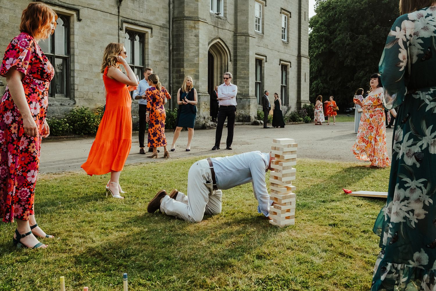 Wedding guest playing Jenga on lawn while others watch outside historic building.