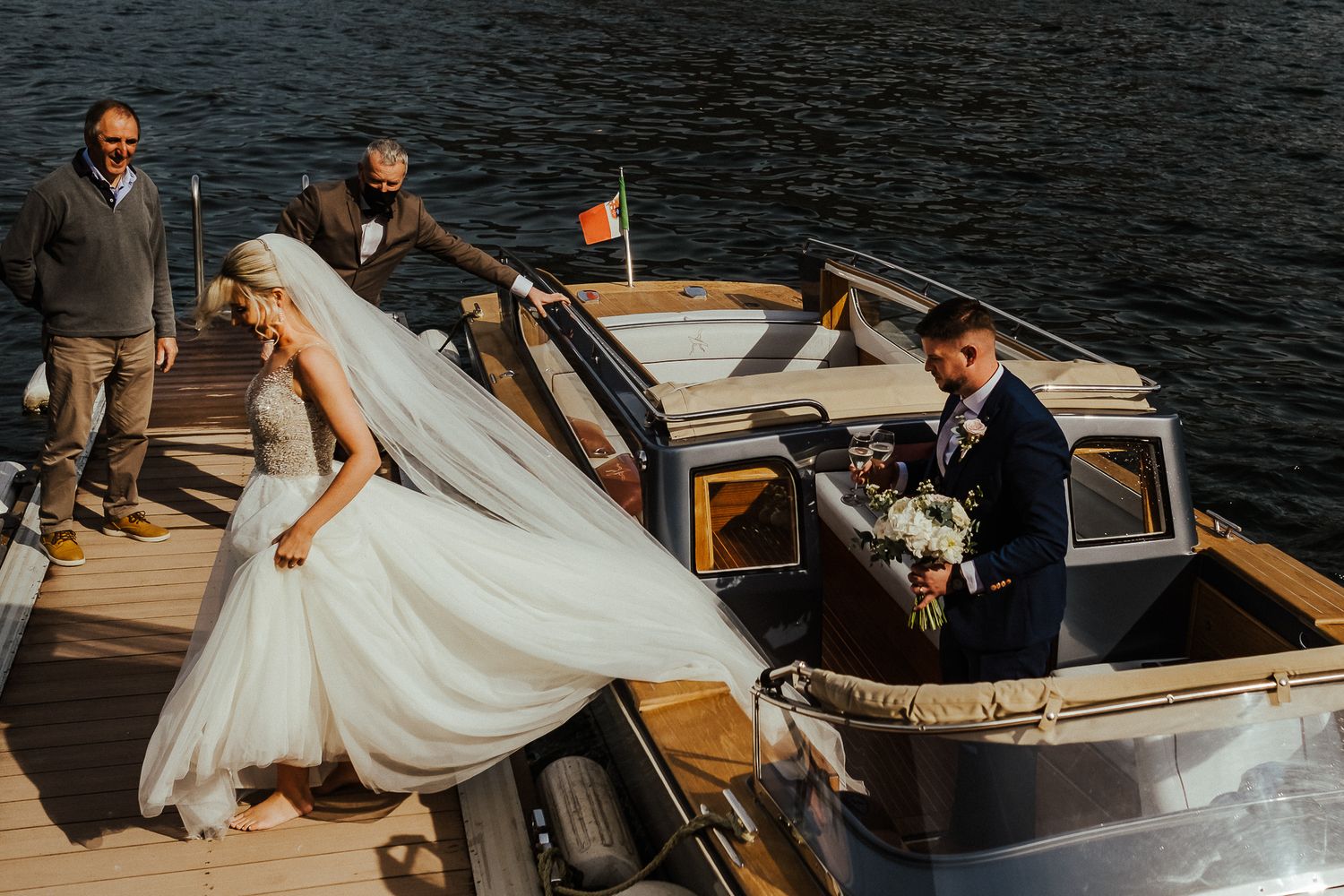 A bride's dress flows in the wind as she steps onto a classic wooden boat.