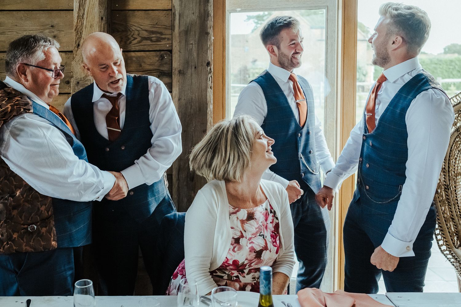 Wedding guests engage in animated conversation around a rustic wooden table.