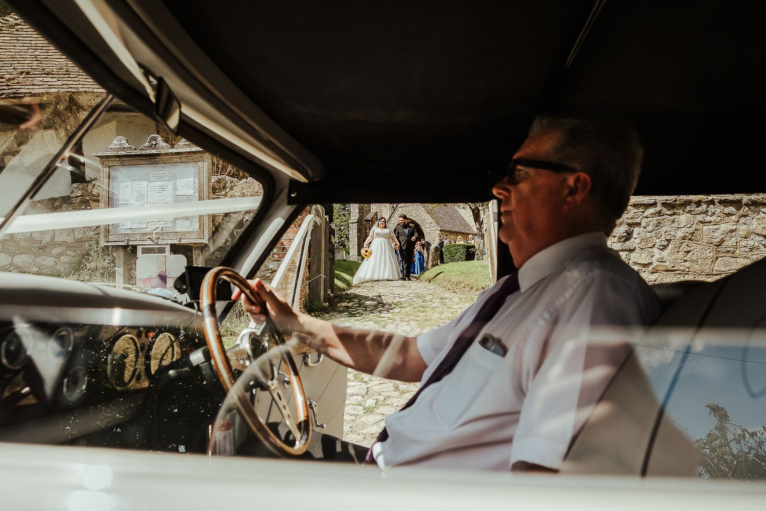 Wedding party members laugh and share drinks while seated in vintage cars.
