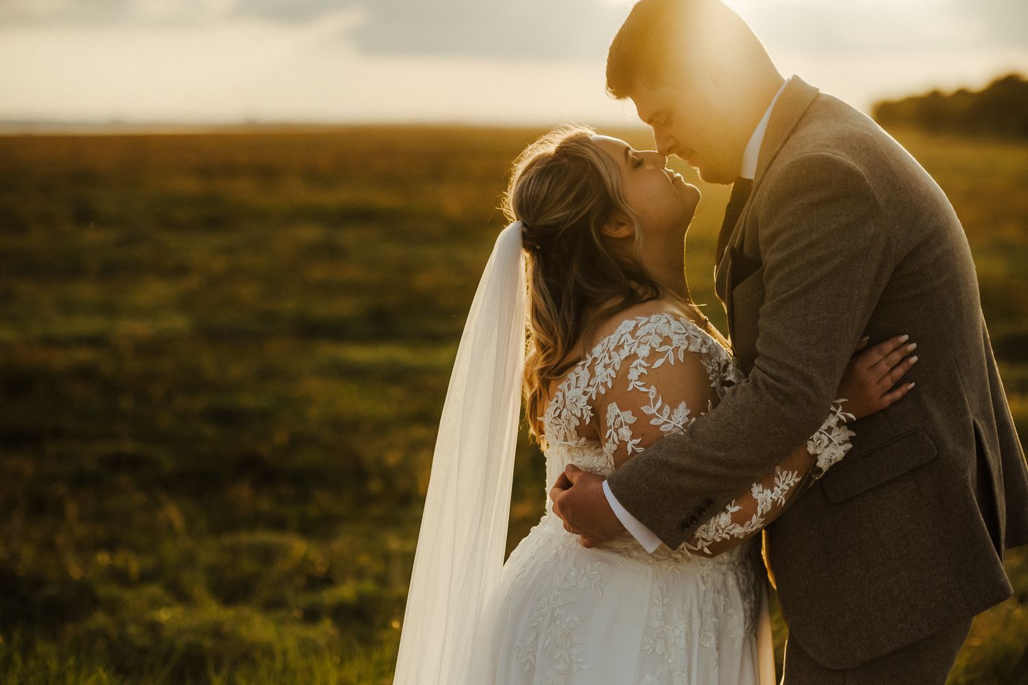 Bride and groom share romantic sunset kiss in open field with golden light.