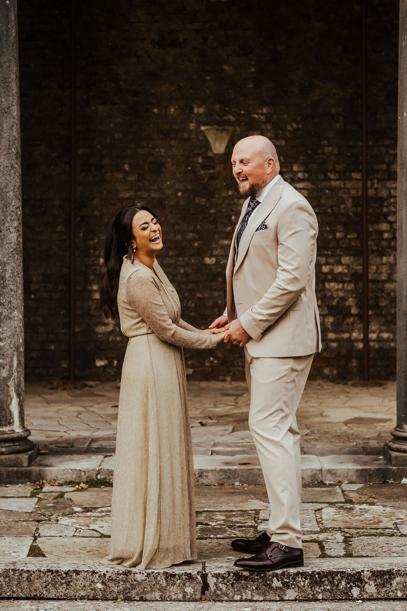 Couple in cream-colored wedding attire holding hands while standing on stone steps against rustic brick wall backdrop.