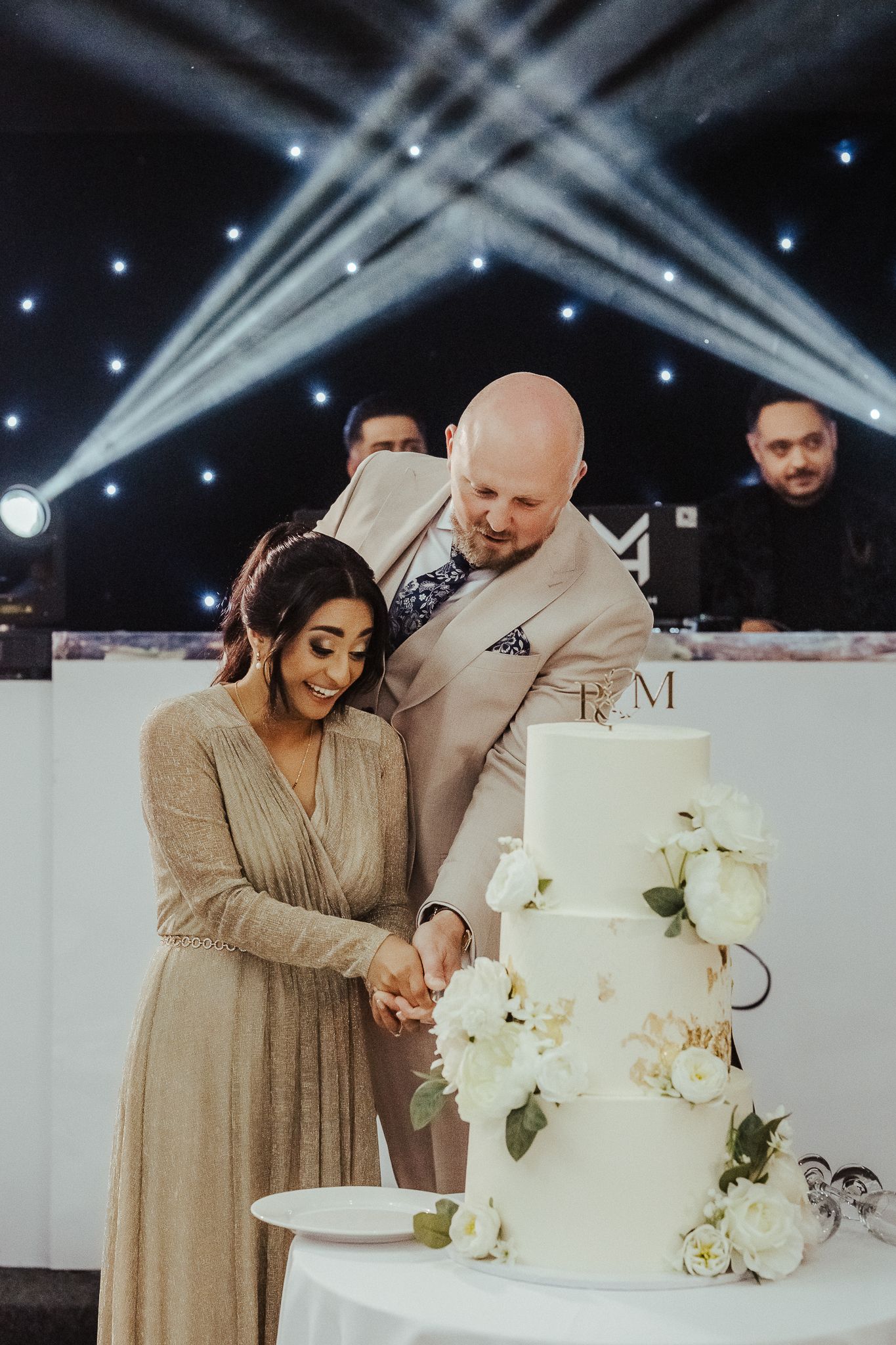 A joyful couple cuts their elegant white wedding cake decorated with white flowers at their reception.