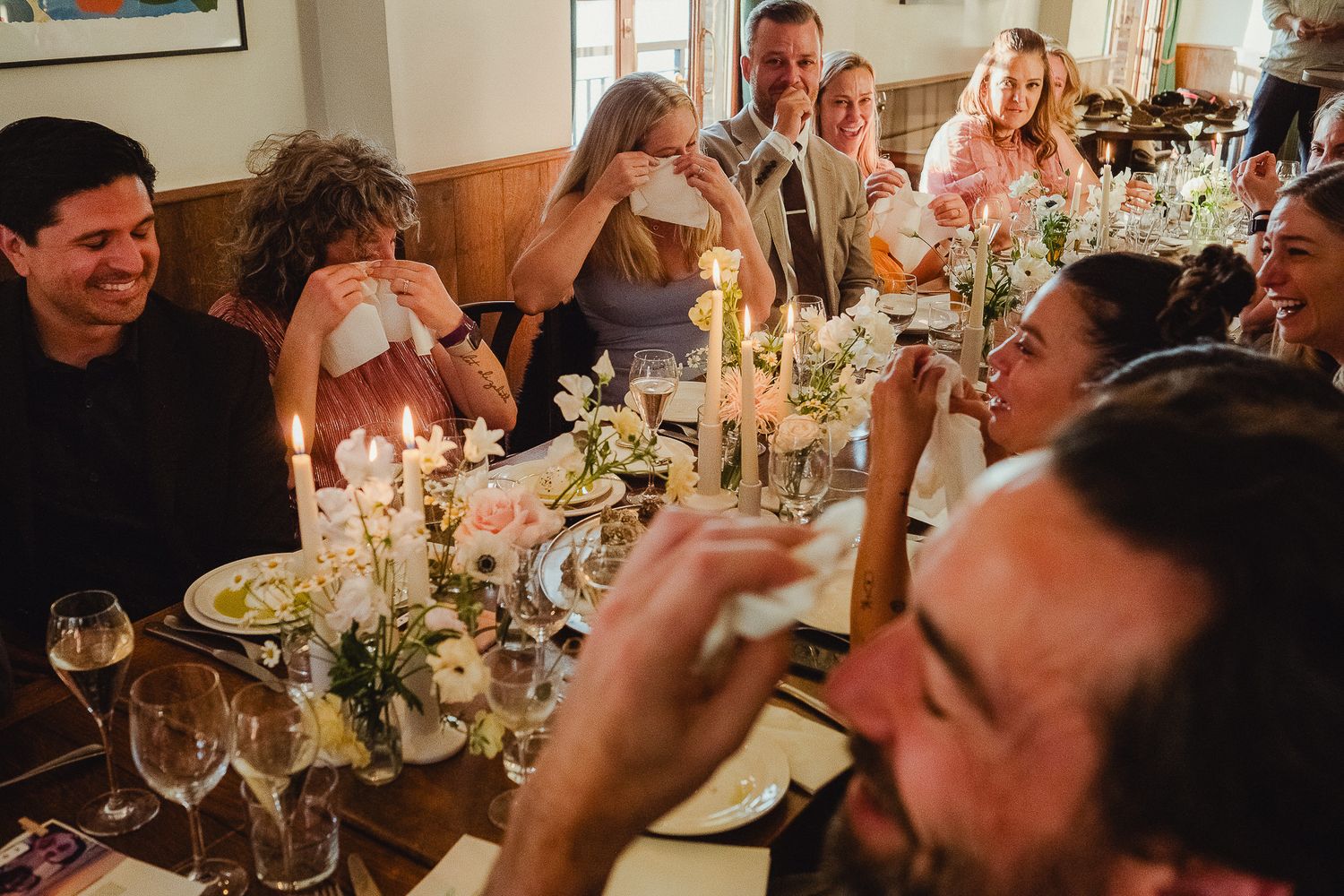 Guests gather around an intimate candlelit dinner table with wine glasses and floral centerpieces for a celebratory meal.