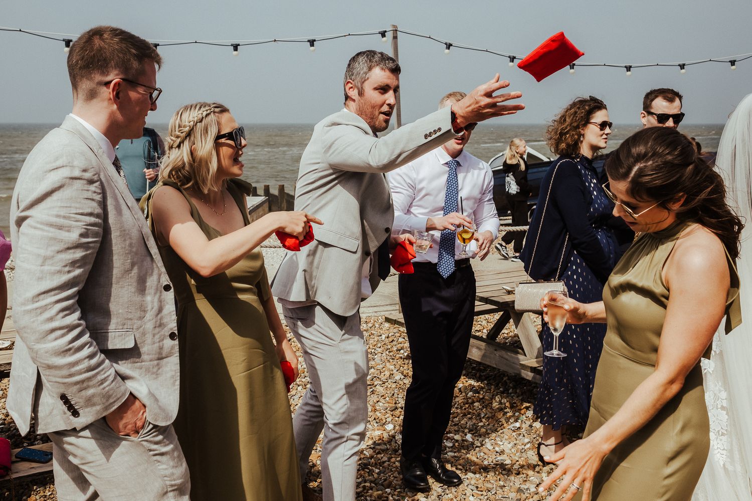 Wedding guests mingle and toast with red drinks at an outdoor beach celebration.