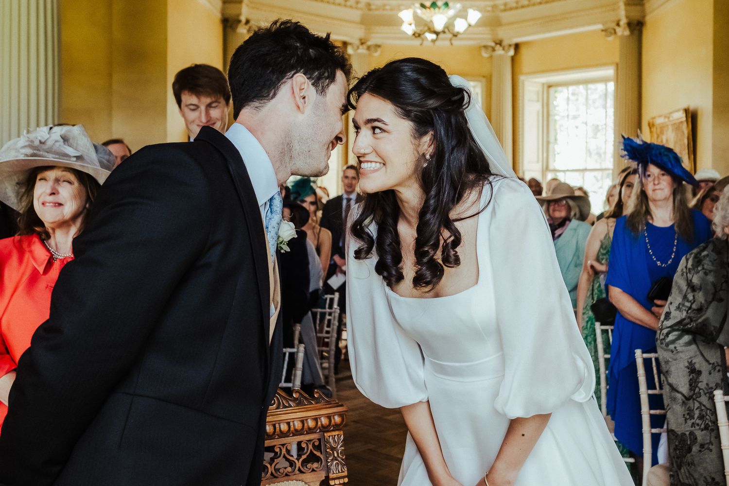 A tender wedding moment between a couple surrounded by guests in an elegant room.