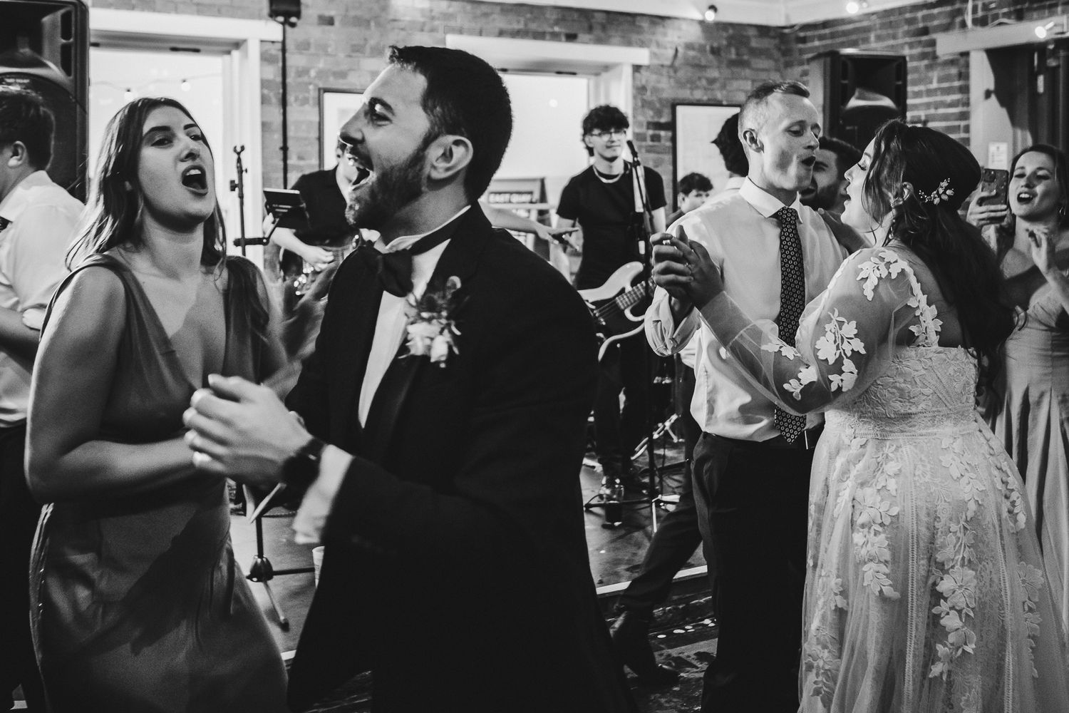 Black and white photo of wedding guests dancing energetically at a rustic indoor reception venue.