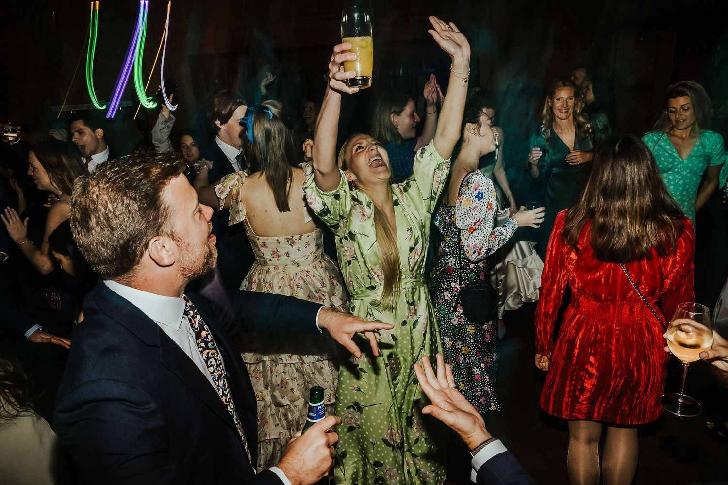 Wedding party celebrates with raised hands and colorful lighting on a dark dance floor.