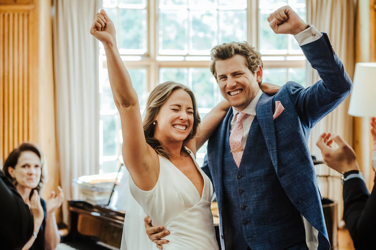 Celebrating newlyweds raise their arms in joy while guests applaud in a sunlit room.